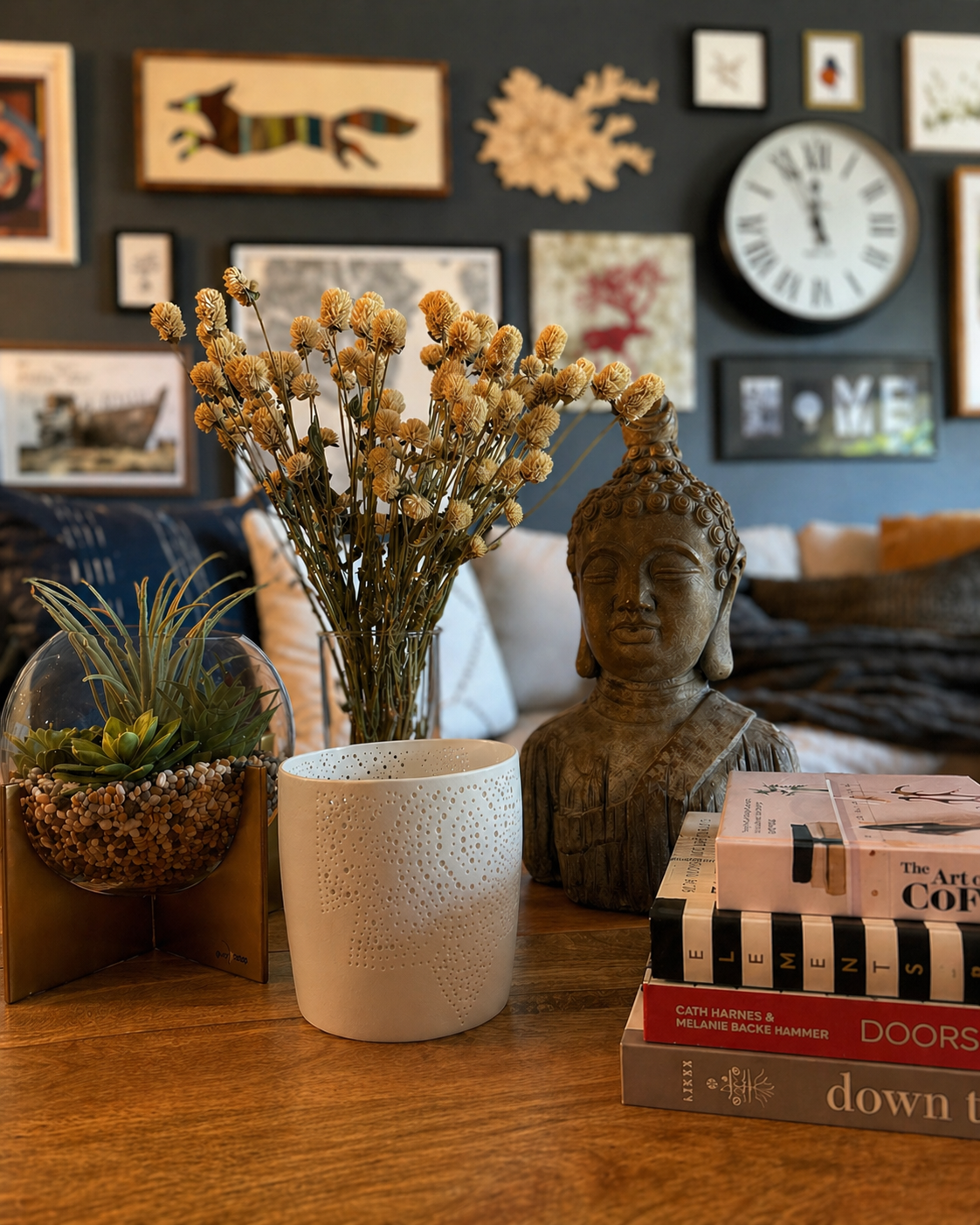 Modern eclectic coffee table styling with dried flowers in a glass vase, a white ceramic candle holder, a sculptural bust, stacked books, and a small terrarium in a cozy living room setting.