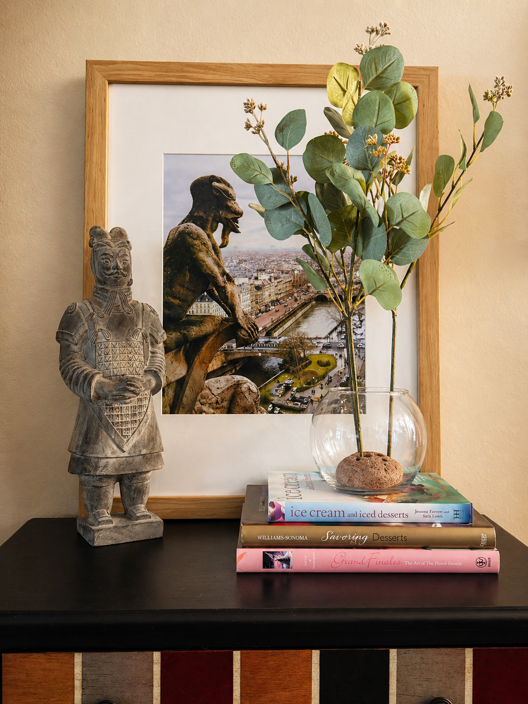 Modern eclectic tabletop vignette with a glass vase holding eucalyptus stems on stacked books, styled with a sculptural figurine and framed artwork in a warm, layered interior.