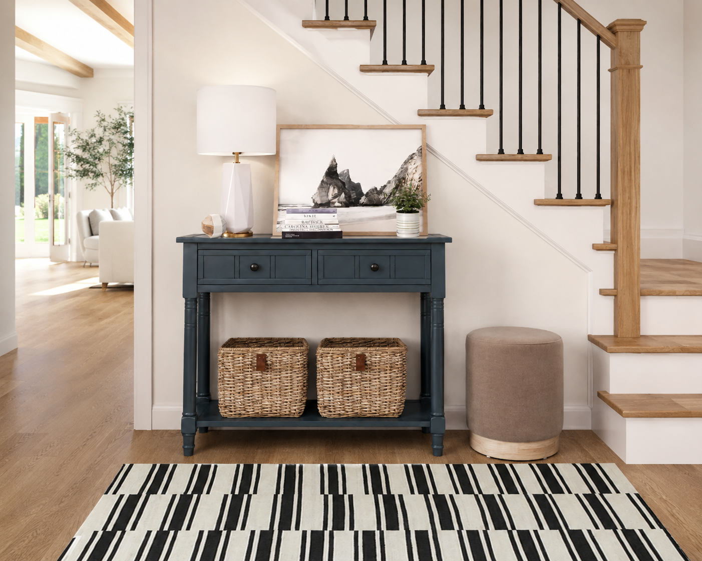 Small foyer with a dark blue console, woven baskets, taupe ottoman, black-and-white rug, coastal artwork, and a wood staircase.