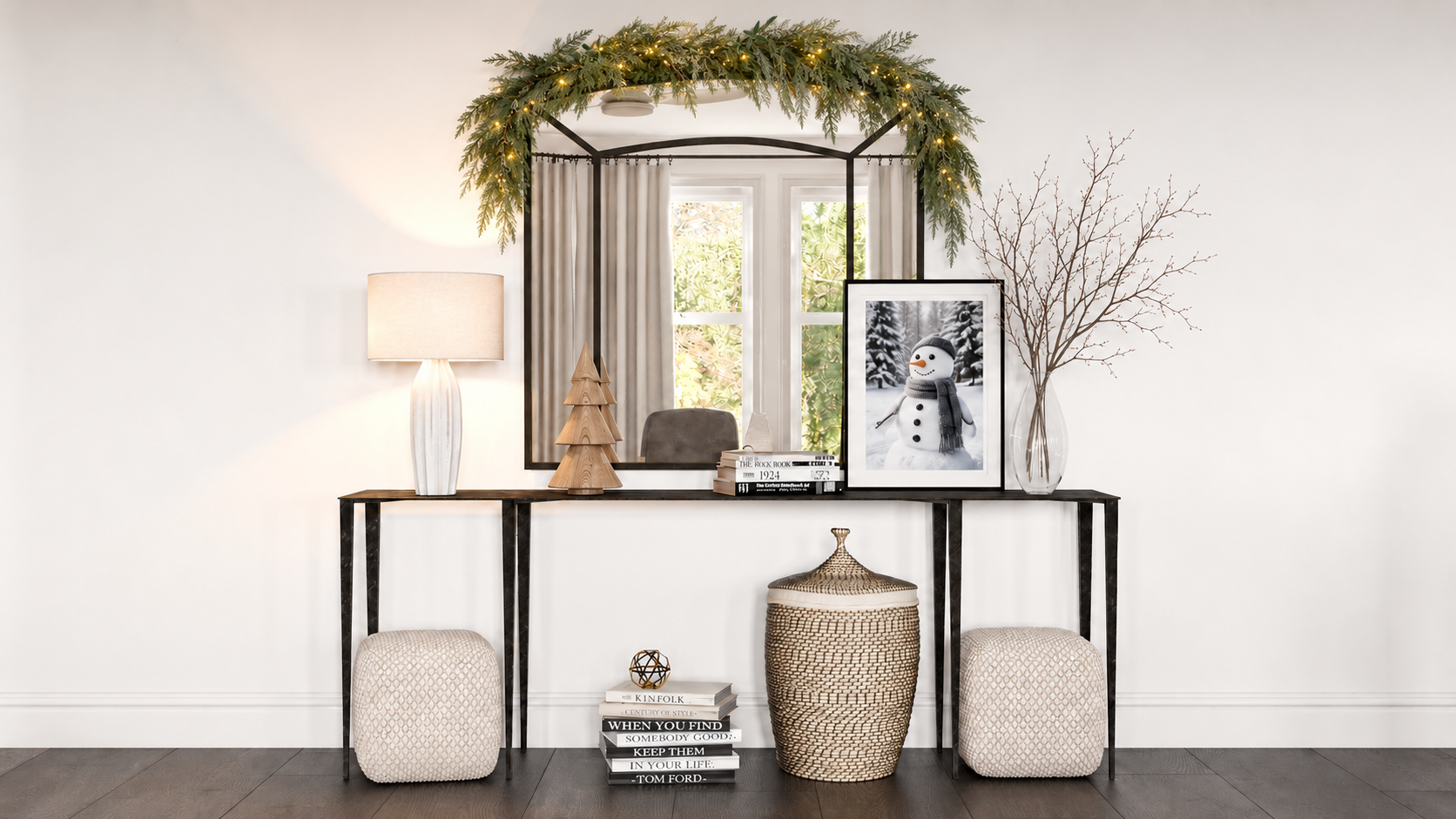 Black-framed mirror topped with lit greenery above a console displaying bare branches in a glass vase, framed snowman photography, and a carved wood Christmas tree.