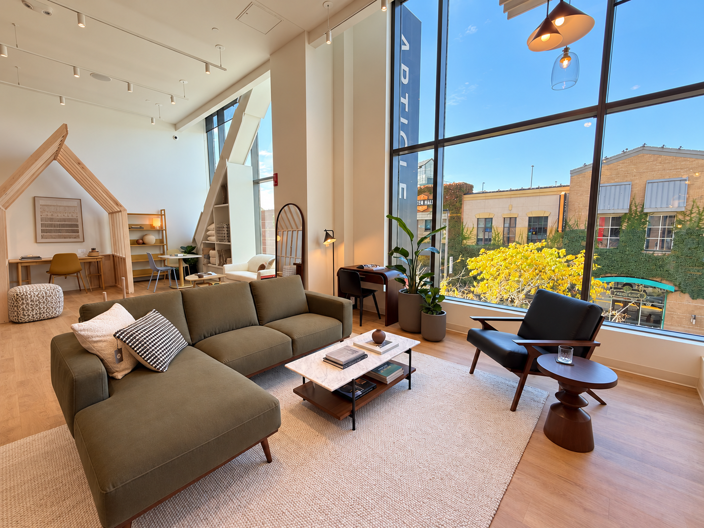 Olive green chaise sectional, black leather lounge chair, marble-top coffee table, and sculptural wood side table arranged on an ivory rug beside tall showroom windows.