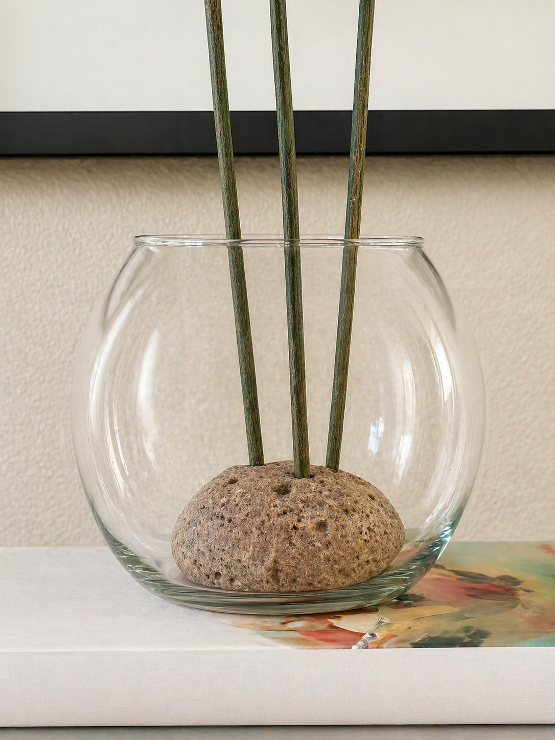 Close-up of a glass vase with a natural stone base and stems, styled on stacked books in a modern eclectic interior with a clean, neutral background.