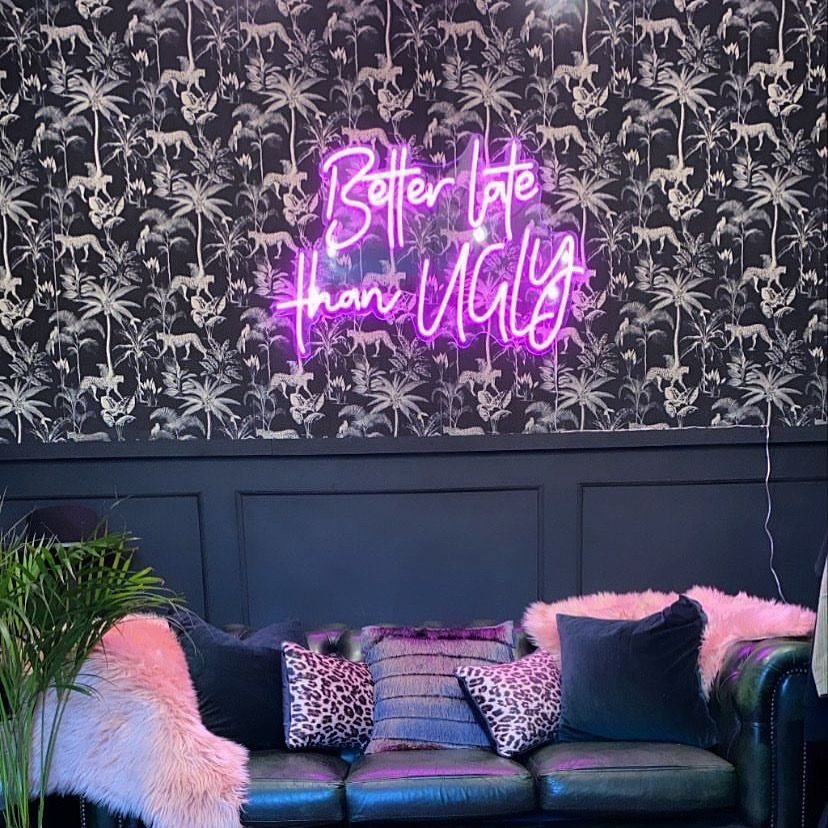 Pink "Better Late Than Ugly" neon sign mounted above a bed in a modern bedroom with botanical wallpaper.