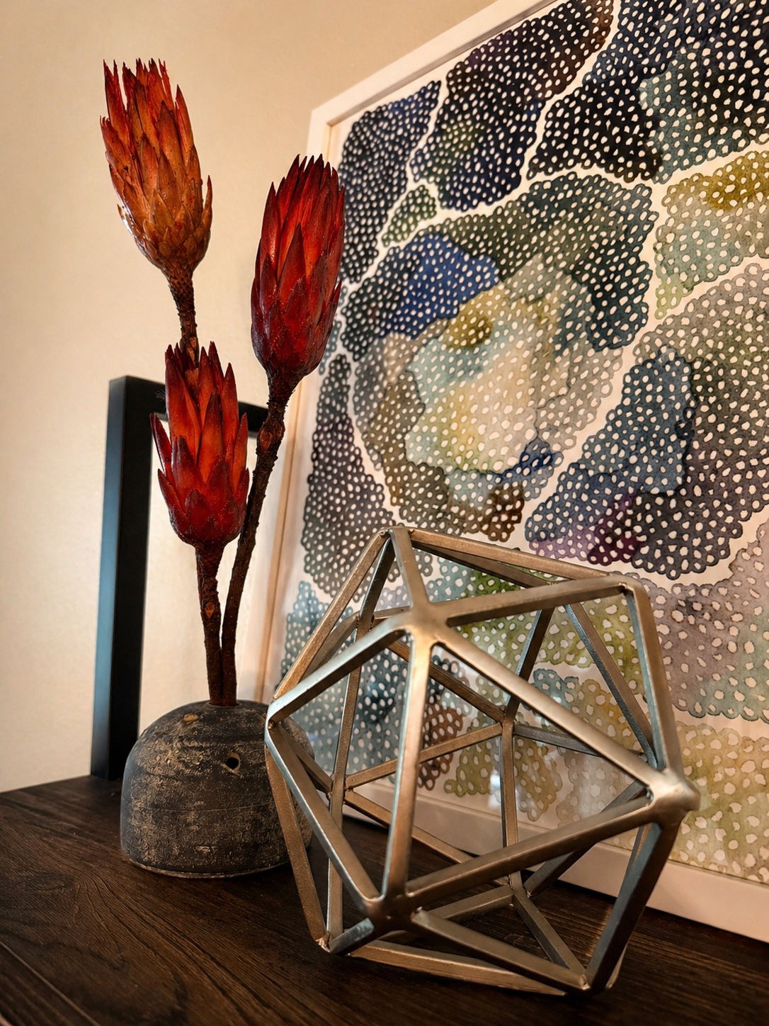 Modern eclectic tabletop vignette featuring red dried stems in a textured vase, a geometric metal sculpture, and layered artwork in a warm, styled interior setting.