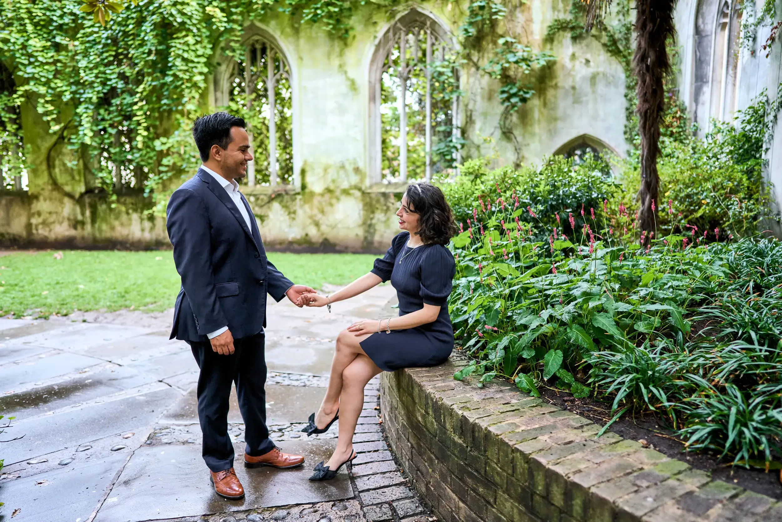 Engaged couple holding hands among ivy-covered Gothic arches at St Dunstan in the East church garden, London