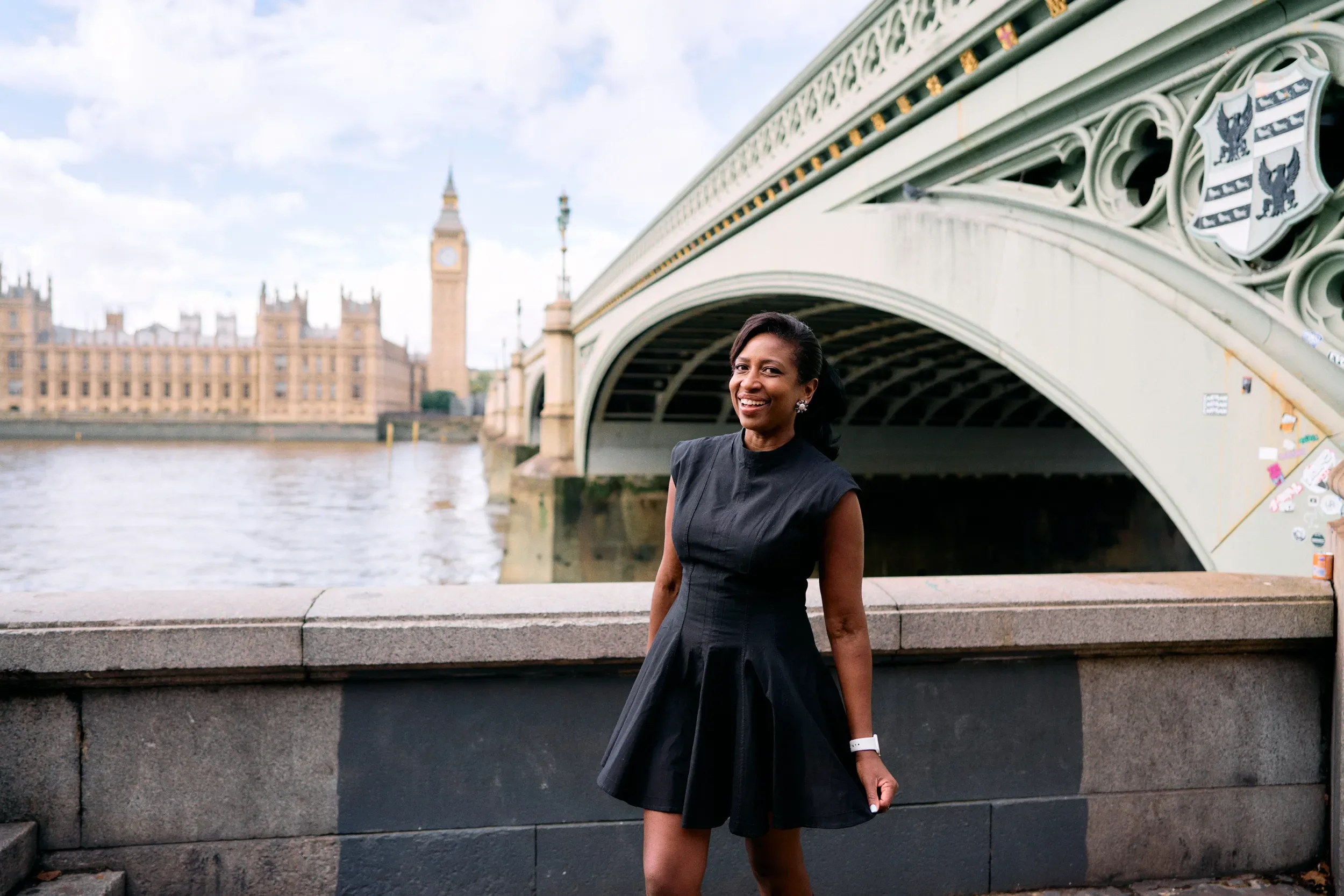 Woman smiling in a black dress on the South Bank with Westminster Bridge, Big Ben and the Houses of Parliament across the Thames during a portrait photoshoot in London
