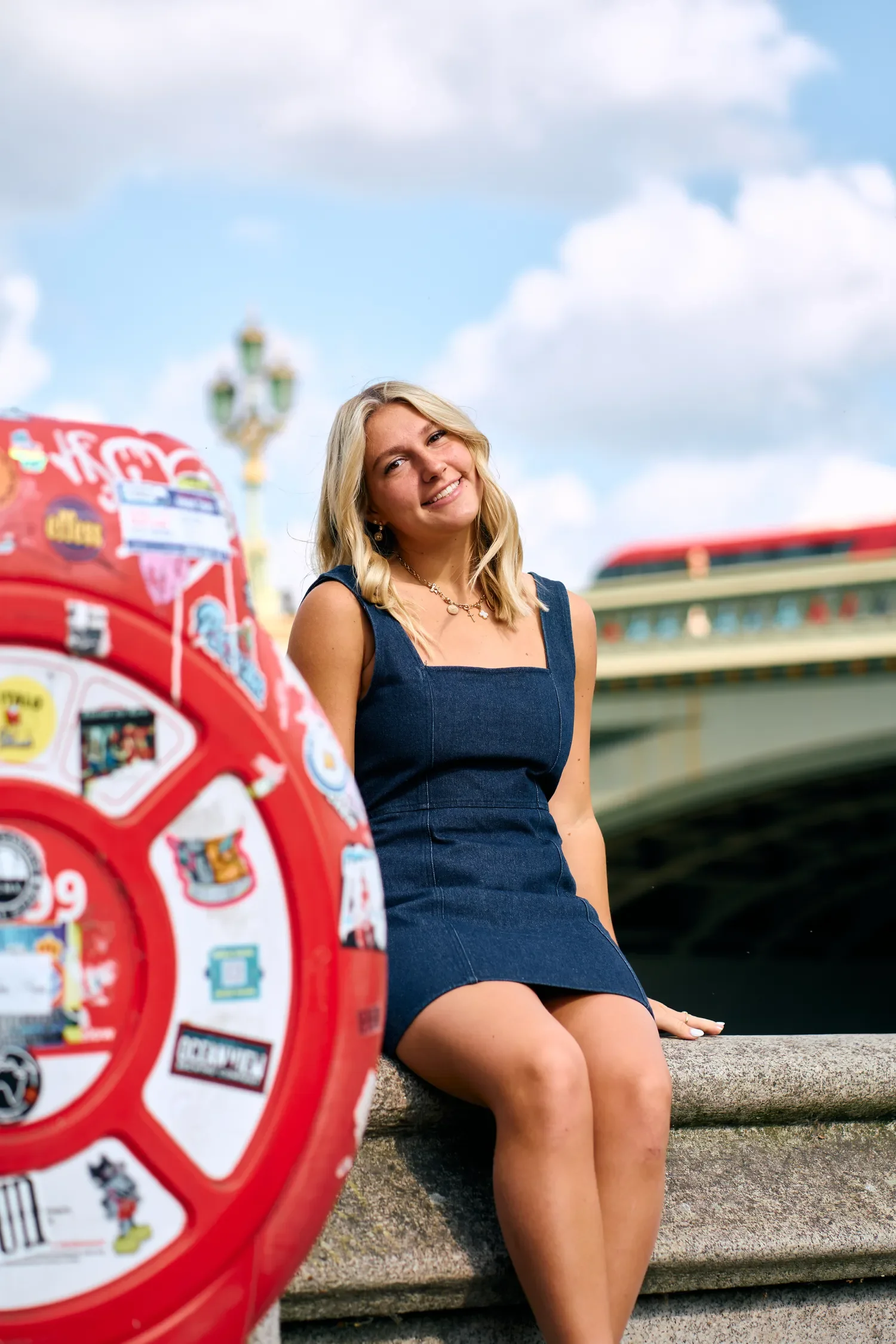 School senior portrait seated beside a sticker-covered red post near Westminster Bridge, with a red London bus behind her