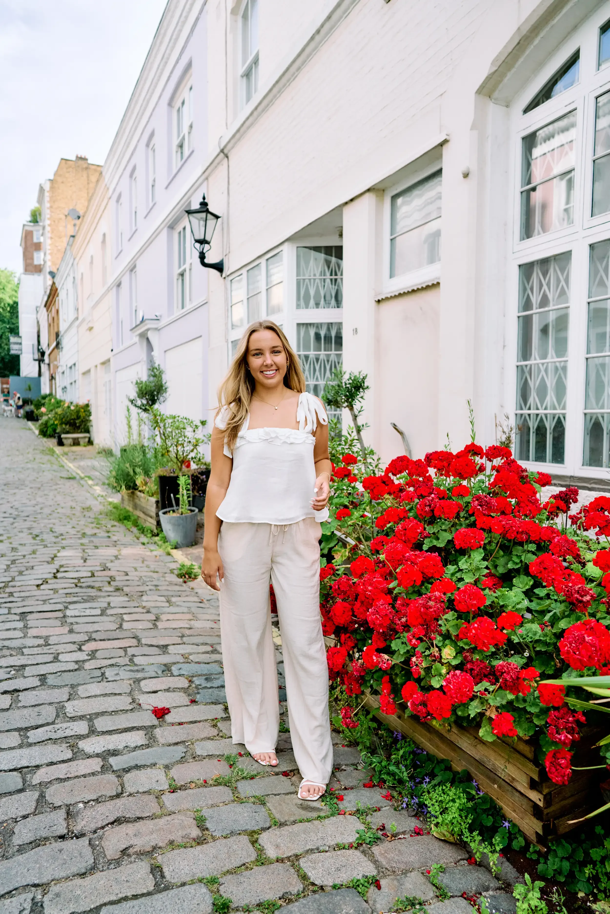 High school senior standing next to bright red geraniums on a cobblestone mews street in Kensington, London