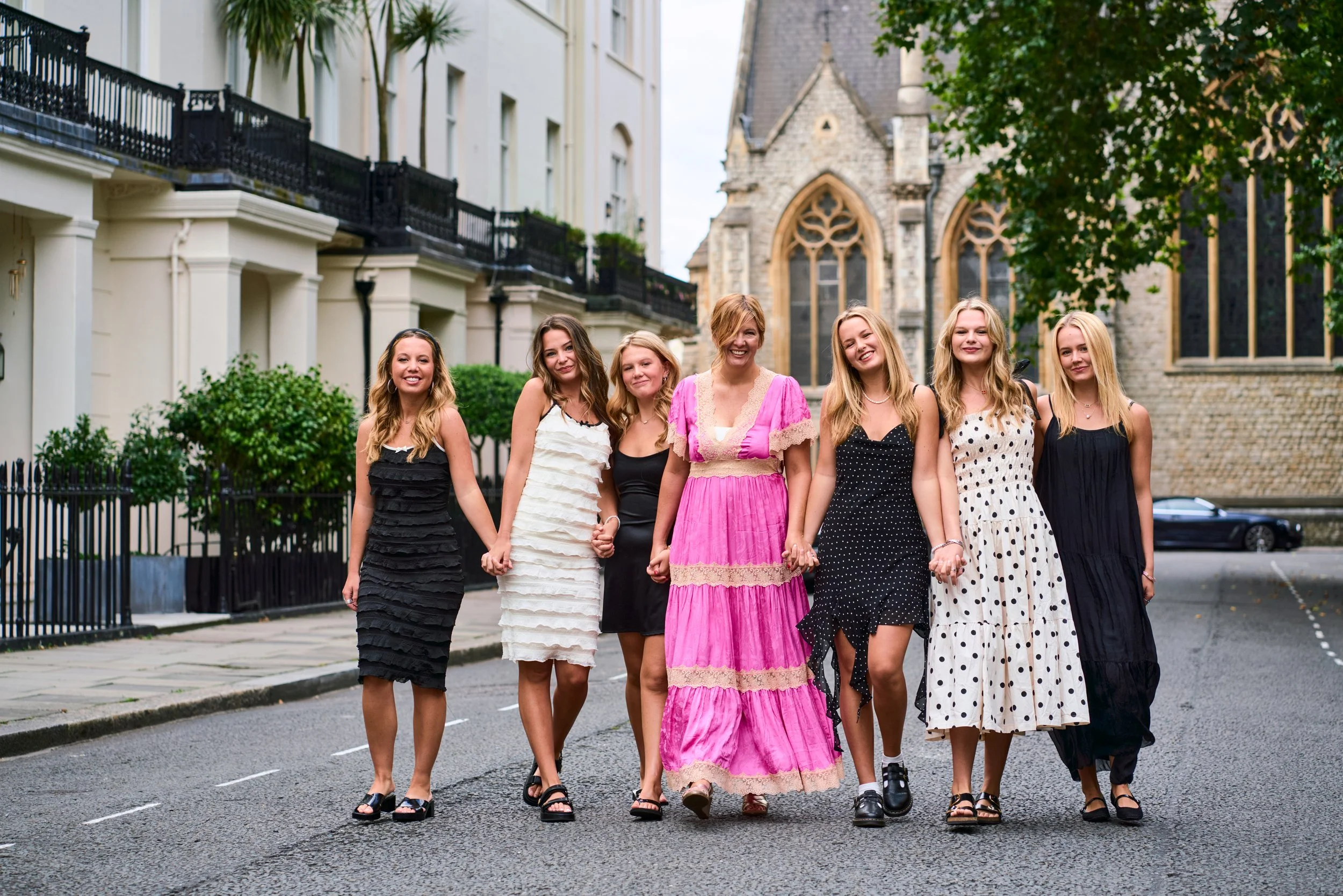 Group of seven women in colourful summer dresses walking hand in hand down a tree-lined Eaton square with a Gothic church in the background — London group photography by Tepewehco
