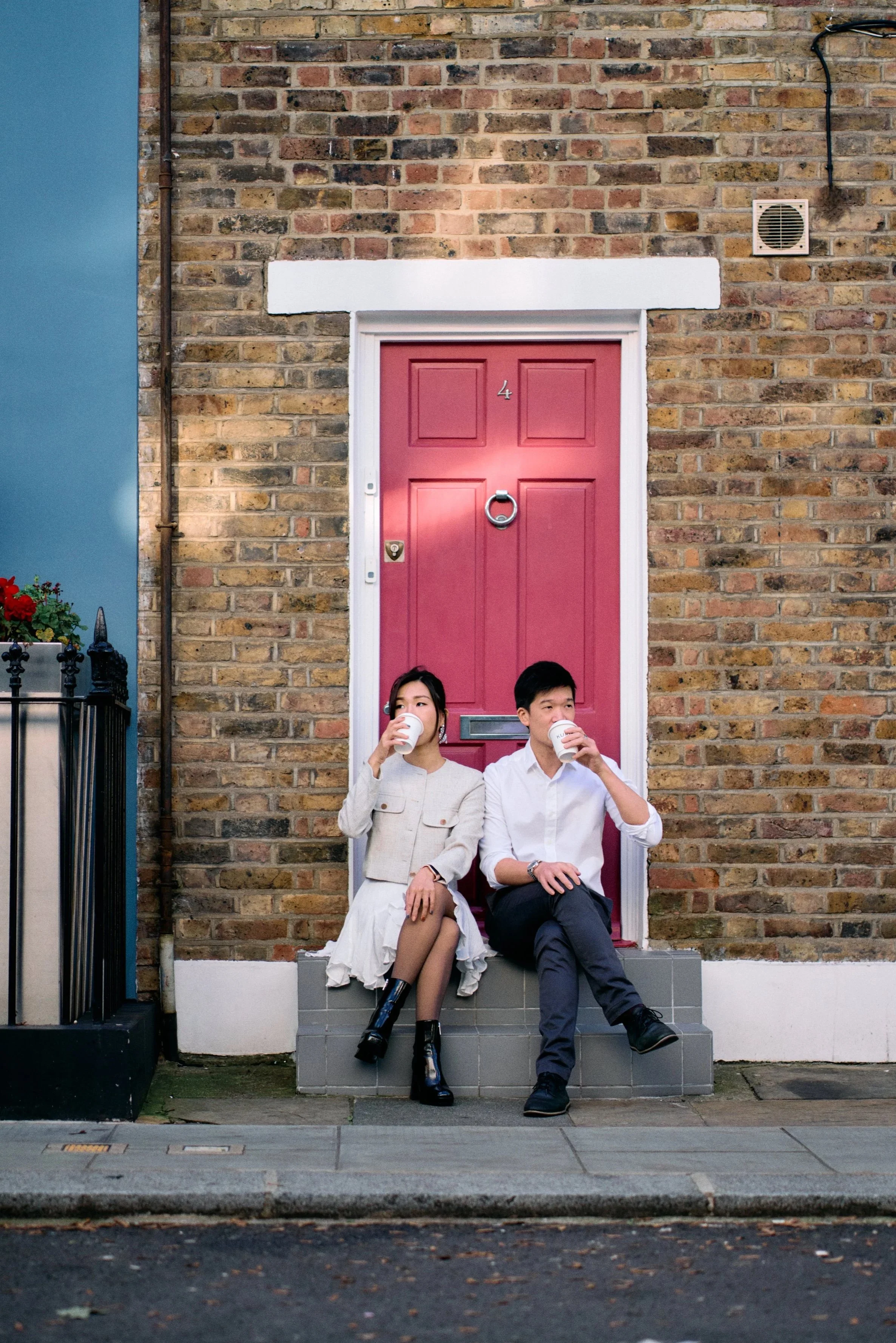 Couple sitting on a doorstep drinking coffee in front of a pink door on a London brick terrace street in Notting Hill