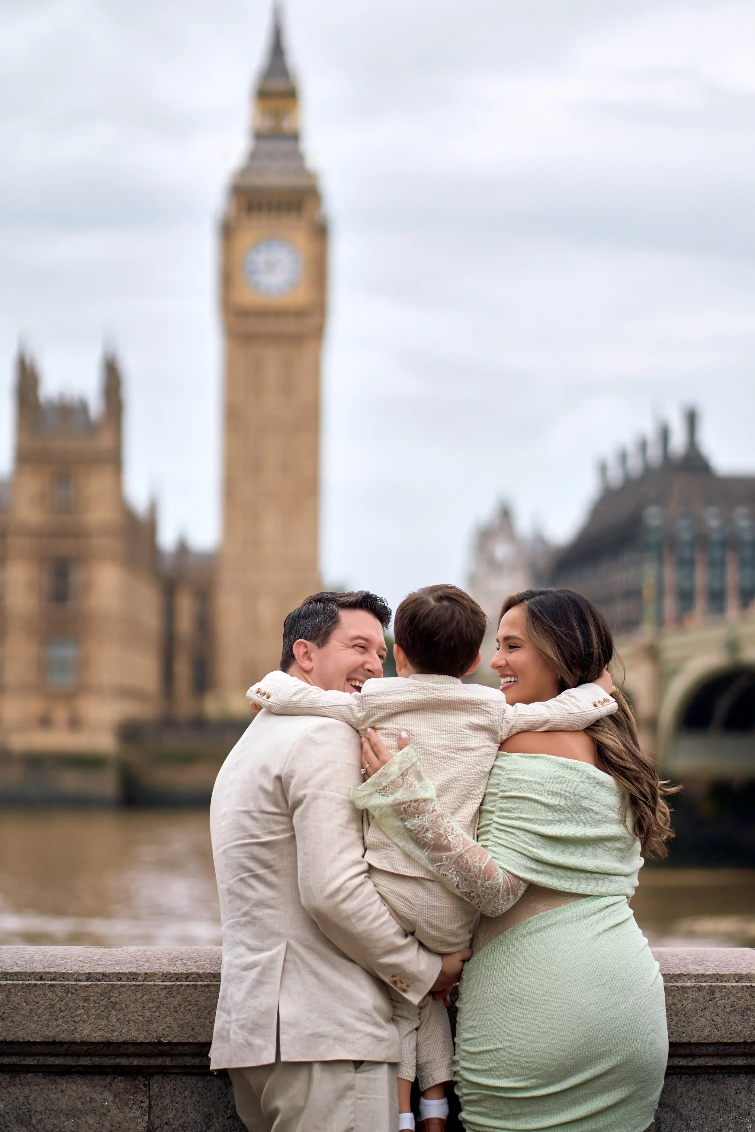 Family of three hugging with their backs to the camera looking towards Big Ben across the Thames during a family photoshoot in London