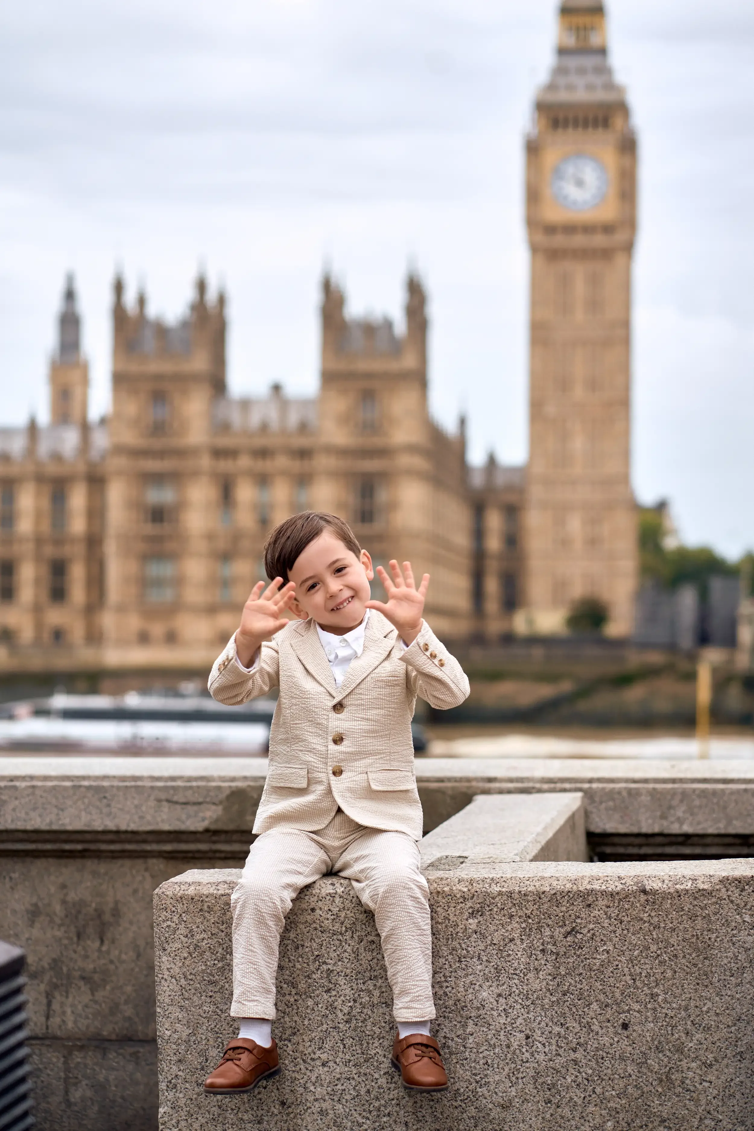 Young boy in a suit sitting on the South Bank wall waving his hands with Big Ben and the Houses of Parliament behind him, London
