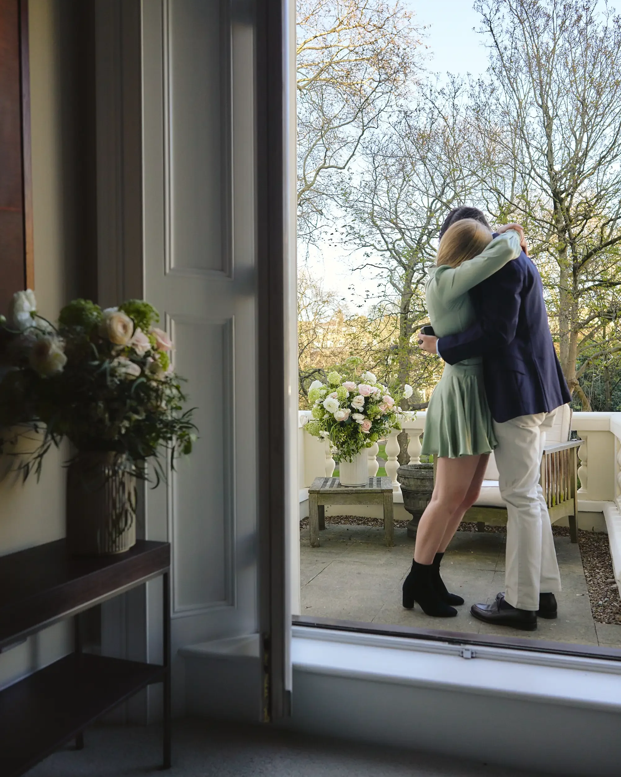 Couple embracing on a flower-decorated terrace framed through an open doorway after a surprise proposal in Chelsea, London