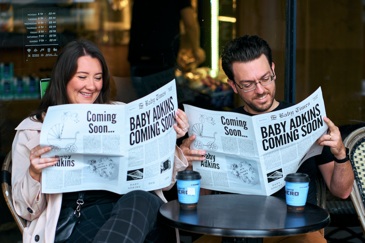 A smiling woman and man sitting at an outdoor cafe table reading newspapers announcing the upcoming arrival of a baby, with two coffee cups on the table.