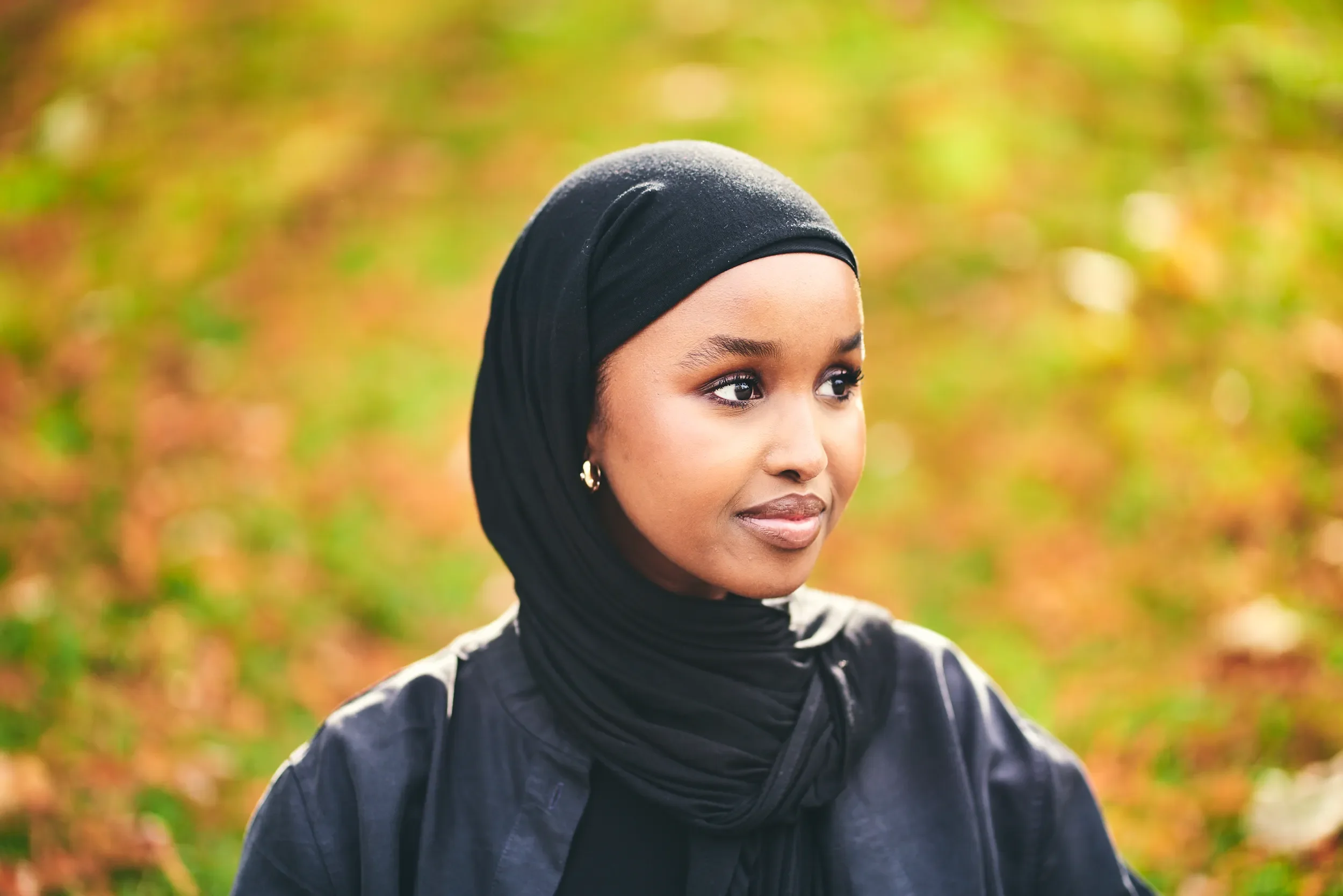 Woman in a black hijab smiling against golden autumn leaves in a London park