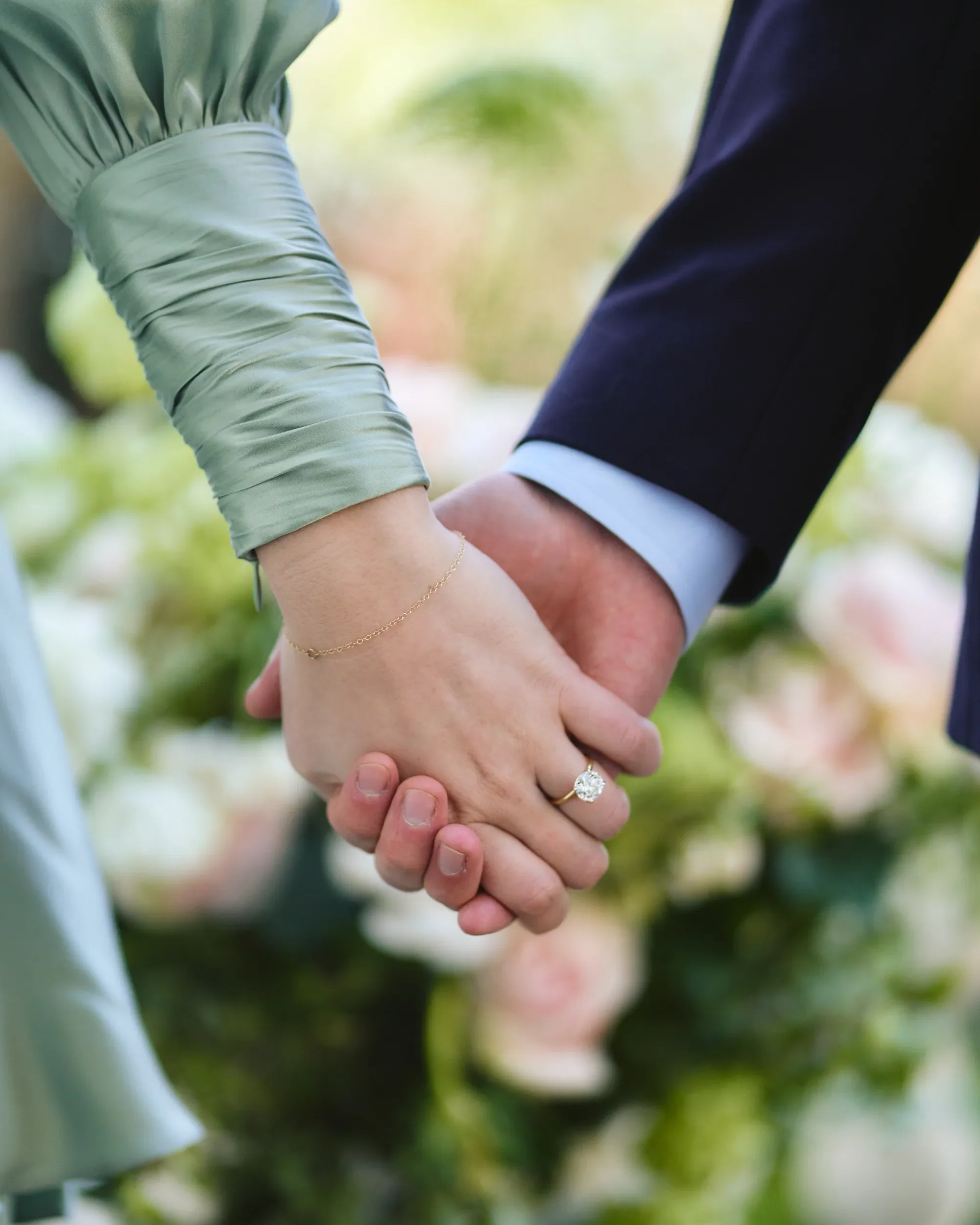 Close-up of an engaged couple holding hands with a diamond ring visible and soft floral bokeh in the background, Chelsea, London