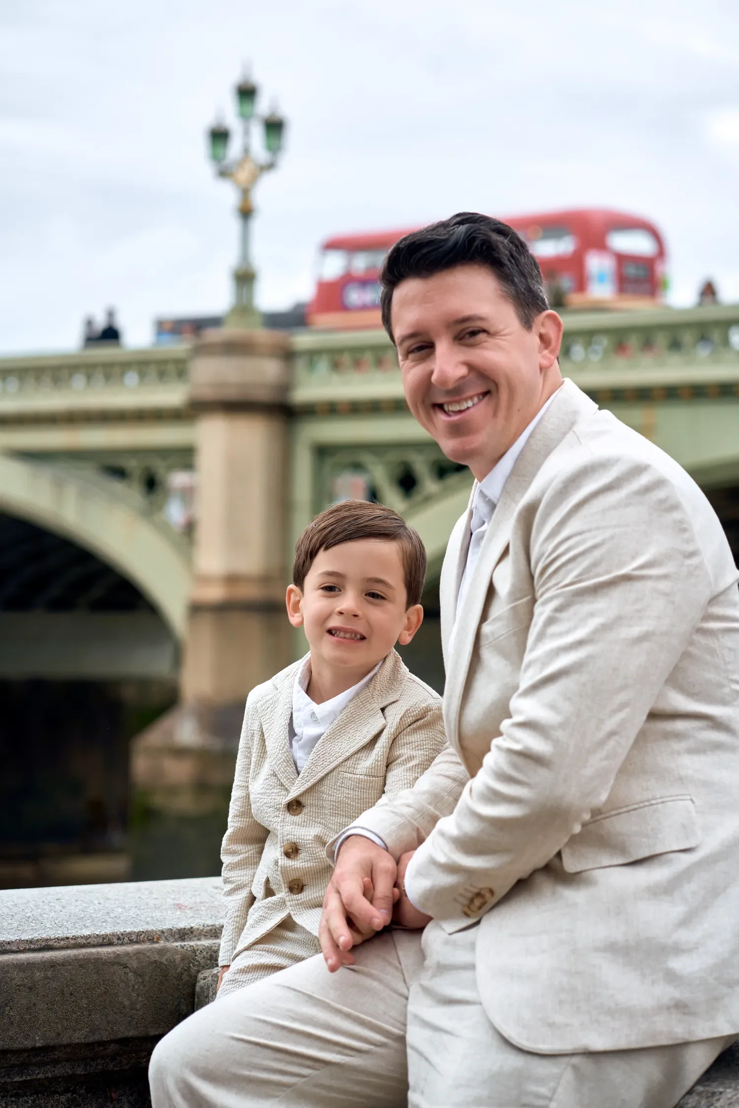 Father and son sitting on Westminster Bridge wall with a red London bus behind
