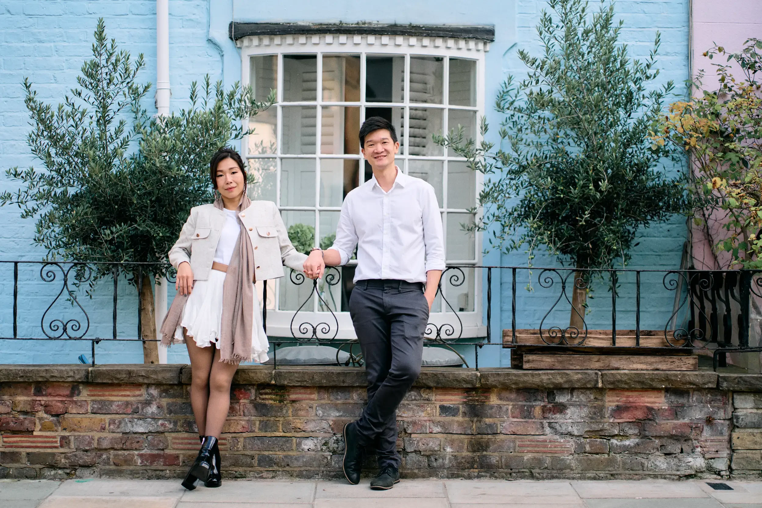 Couple holding hands leaning against a baby blue painted house with olive trees and wrought iron railings in Notting Hill, London - Tepewehco Photography by Kate