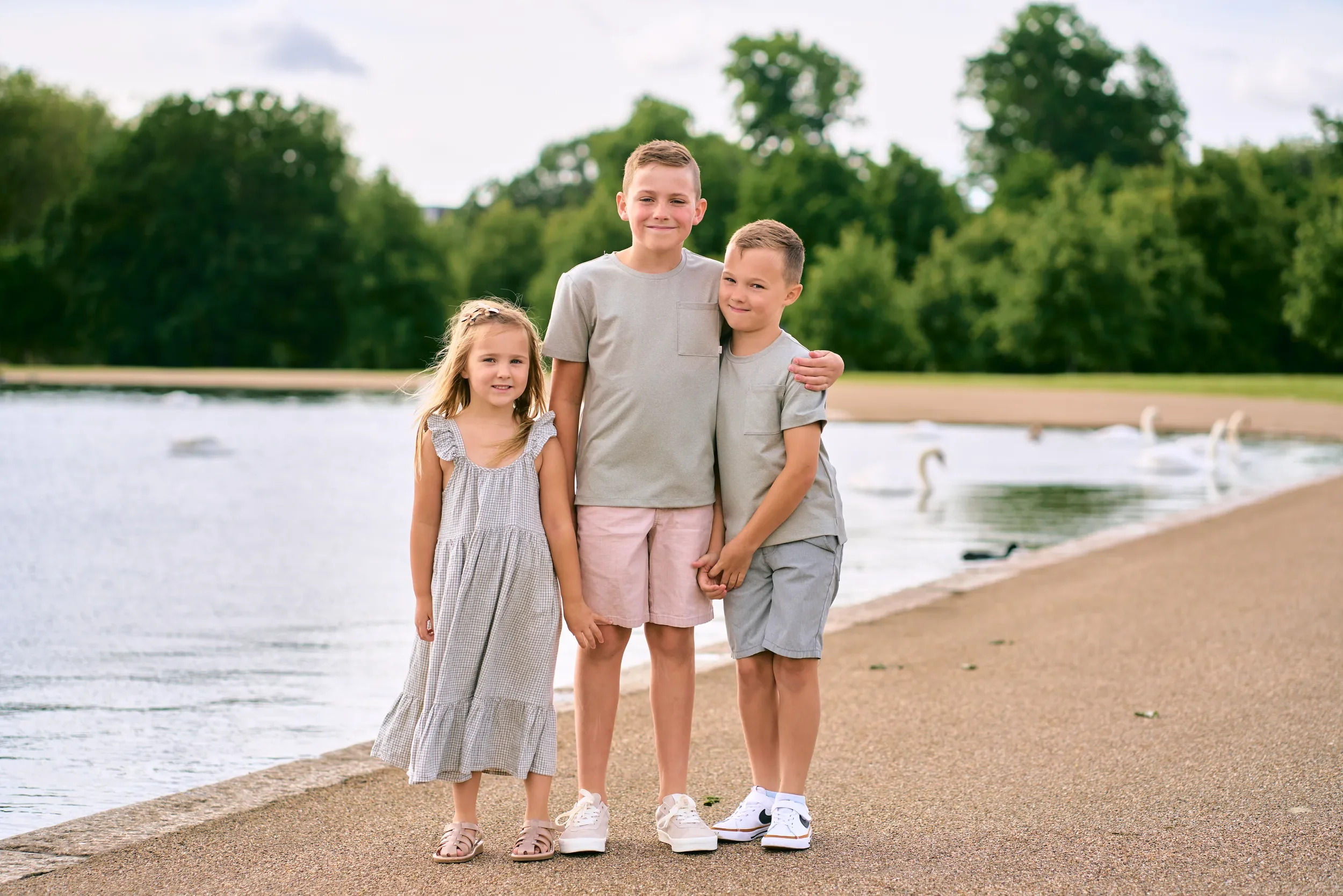 Three siblings in coordinating soft grey and pastel outfits posing by the Serpentine with swans in the background, London — what to wear for a family photoshoot
