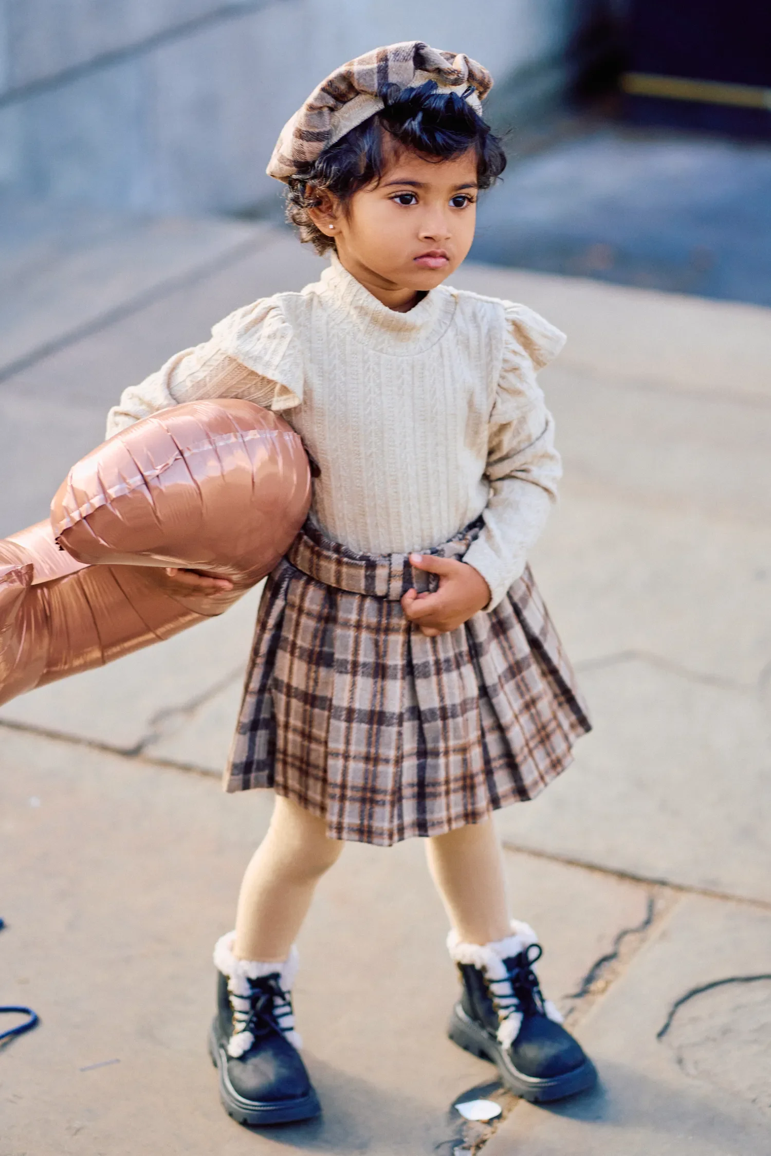 Toddler girl in a plaid skirt and beret holding a rose gold balloon