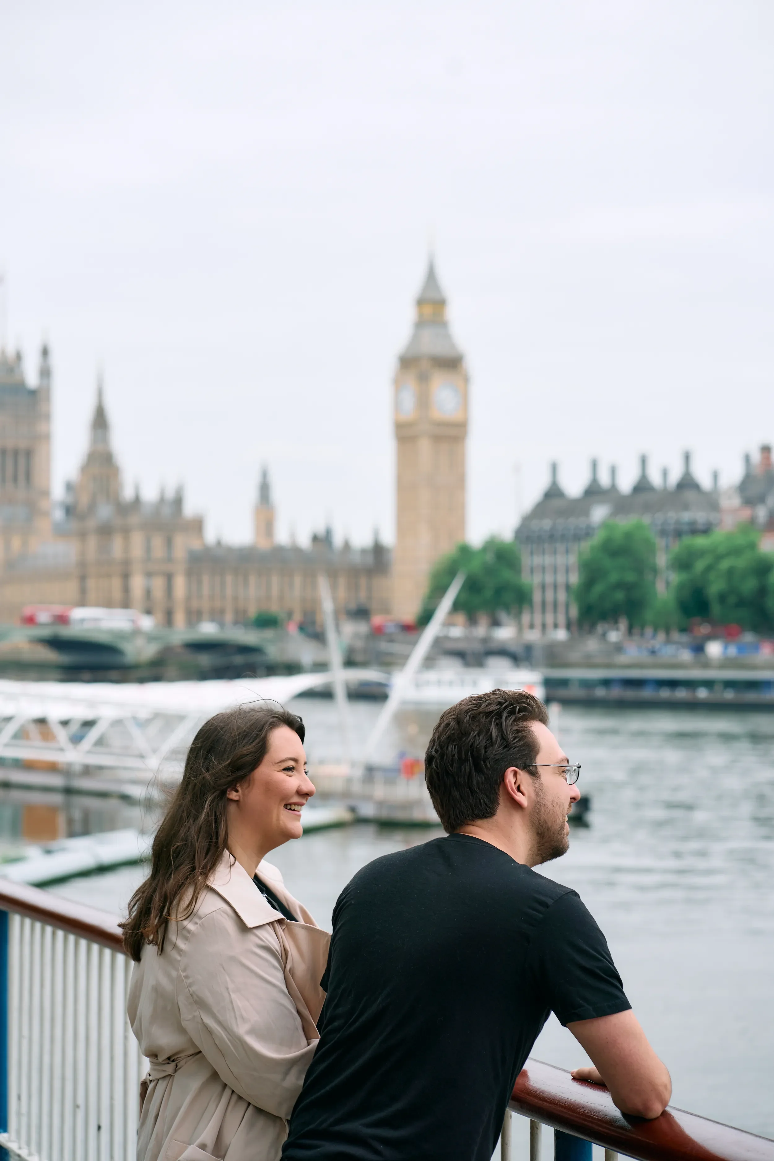 Couple smiling at each other leaning on a railing with Big Ben and river Thames in the background during a couples photoshoot in London