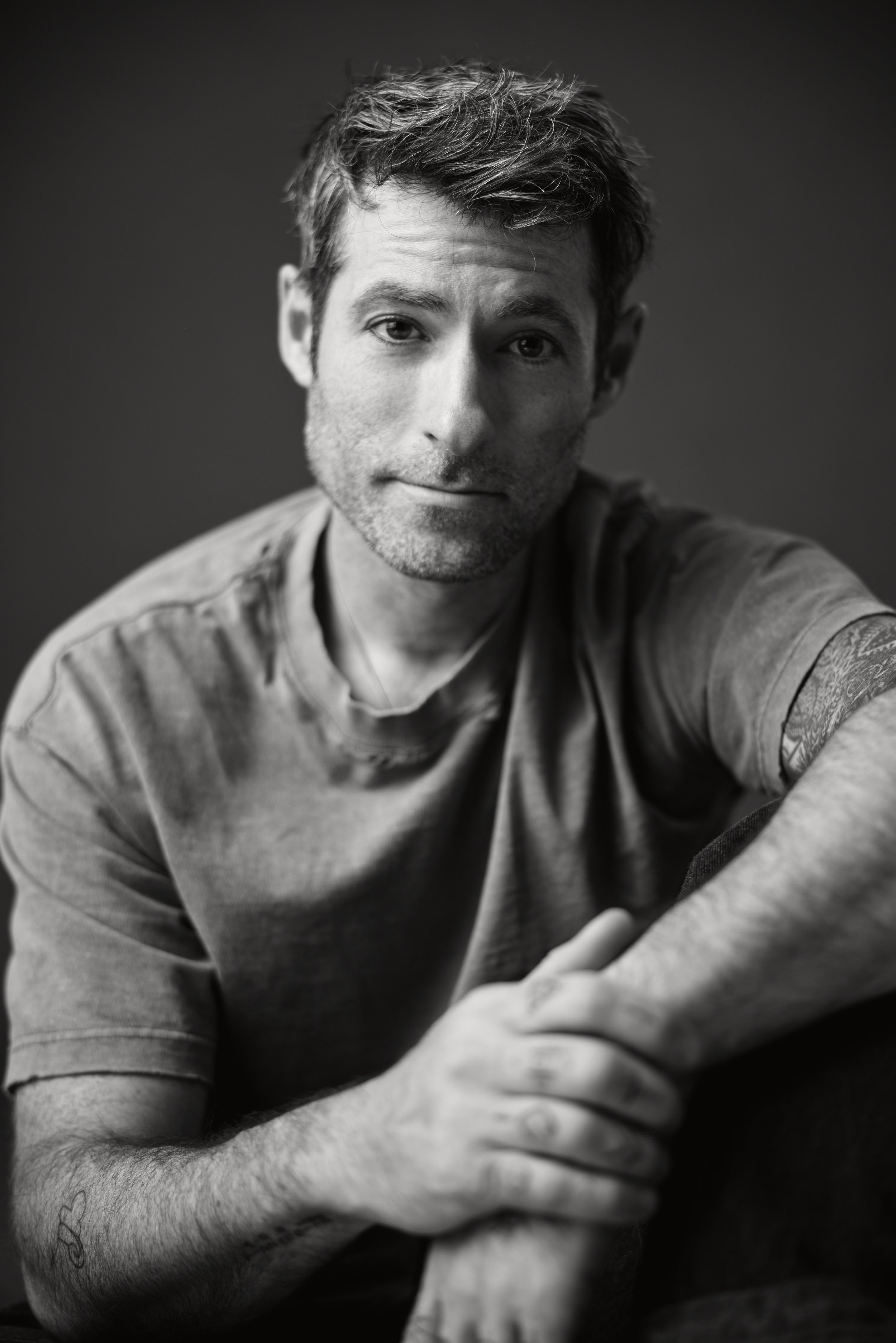 Black and white close-up photo of a young man with textured hair and stubble, wearing a casual t-shirt, looking directly into the camera with a relaxed expression, against a dark plain background.