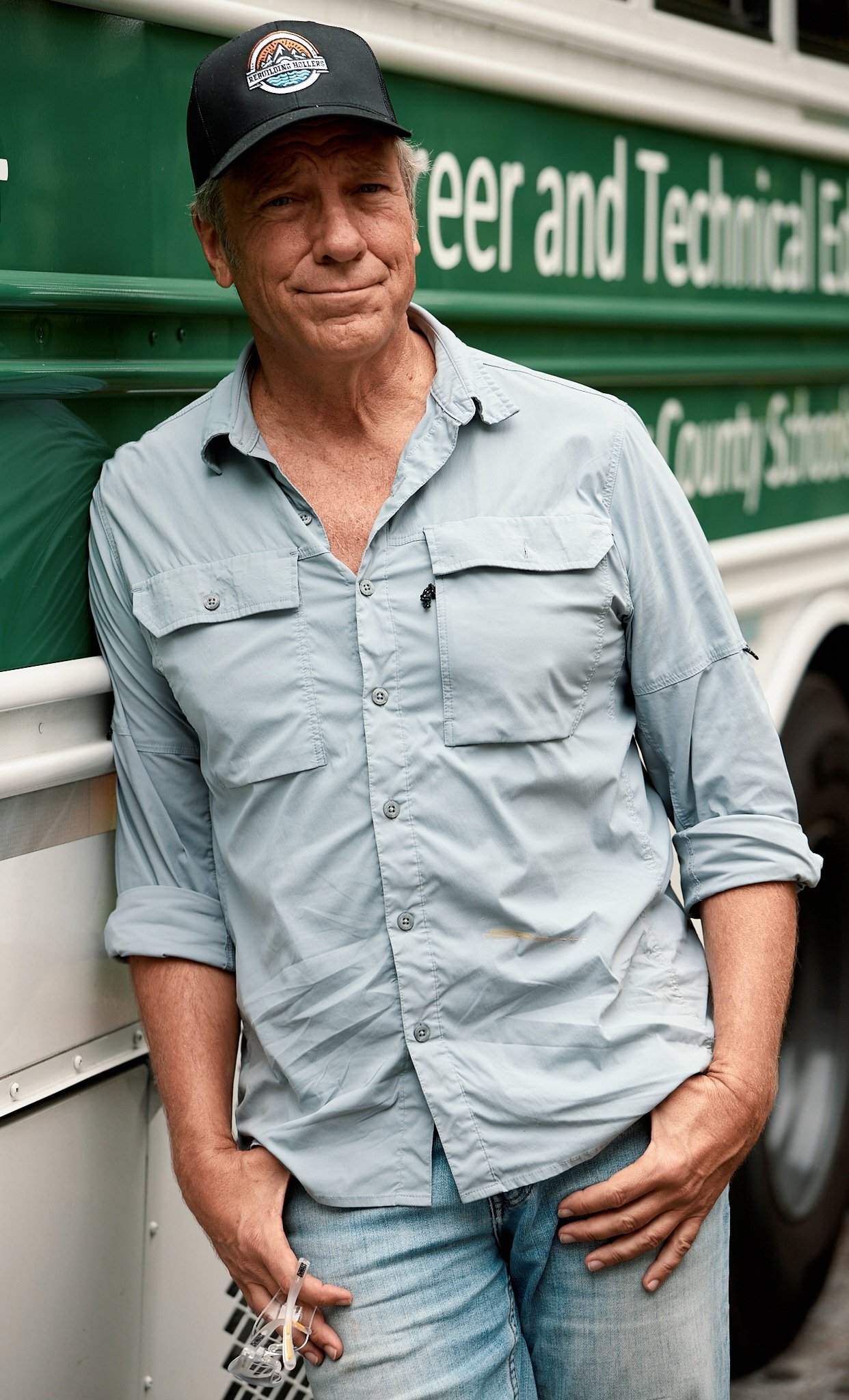 A middle-aged man wearing a black baseball cap, light gray button-up shirt with rolled-up sleeves, and blue jeans. He is standing outdoors next to a green vehicle with white text on it, holding a face mask with glasses in one hand and resting his oth