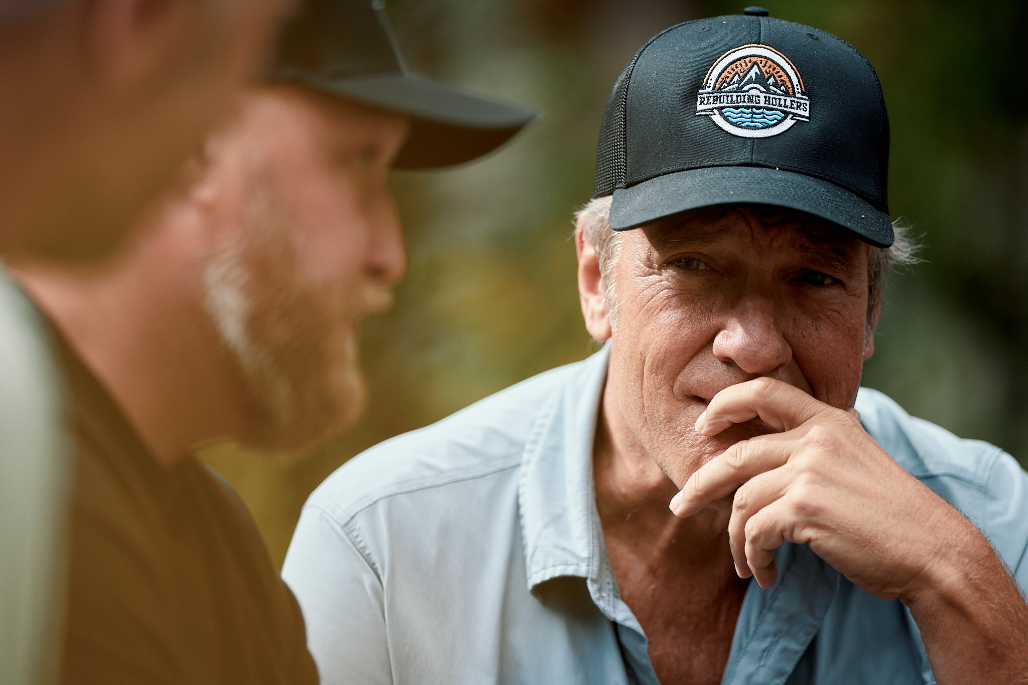 Two men in a serious conversation outdoors, one wearing a black cap with a logo that says 'Rebuilding Rollers' and a light blue shirt, the other with a blurred face wearing a green baseball cap.
