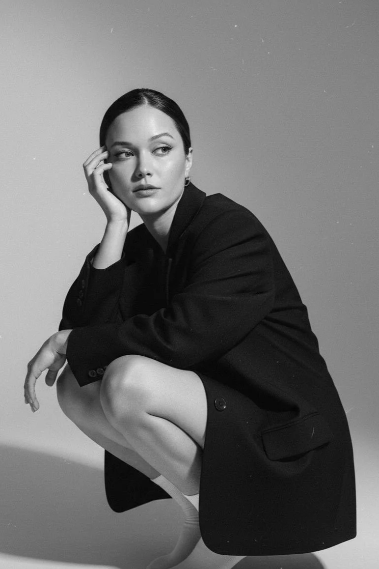 A black and white photo of a woman squatting with her right elbow on her knee and her hand resting on her face. She has dark hair pulled back and is wearing a dark blazer and socks.