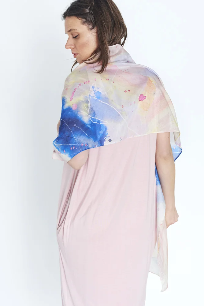 WOMAN IN A PINK DRESS LOOKING OVER HER SHOULDER WEARING A MULTI-COLOURED COTTON SILK SCARF WITH ABSTRACT PAINTERLY PATTERN IN LIGHT YELLOW, PINK AND BLUE OVER HER SHOULDERS. 