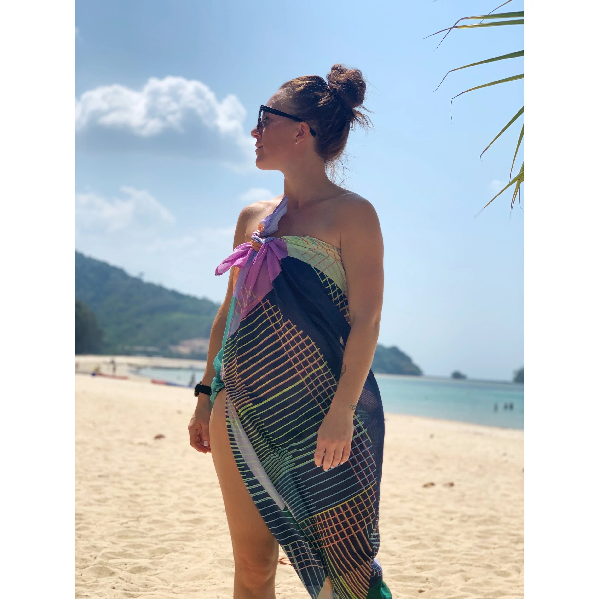 WOMAN ON THE BEACH WEARING A SQUARE COTTON SILK SCARF WITH A GEOMETRIC DESIGN IN BLUE, GREEN ORANGE AND PINK AS A BEACH SARONG. 