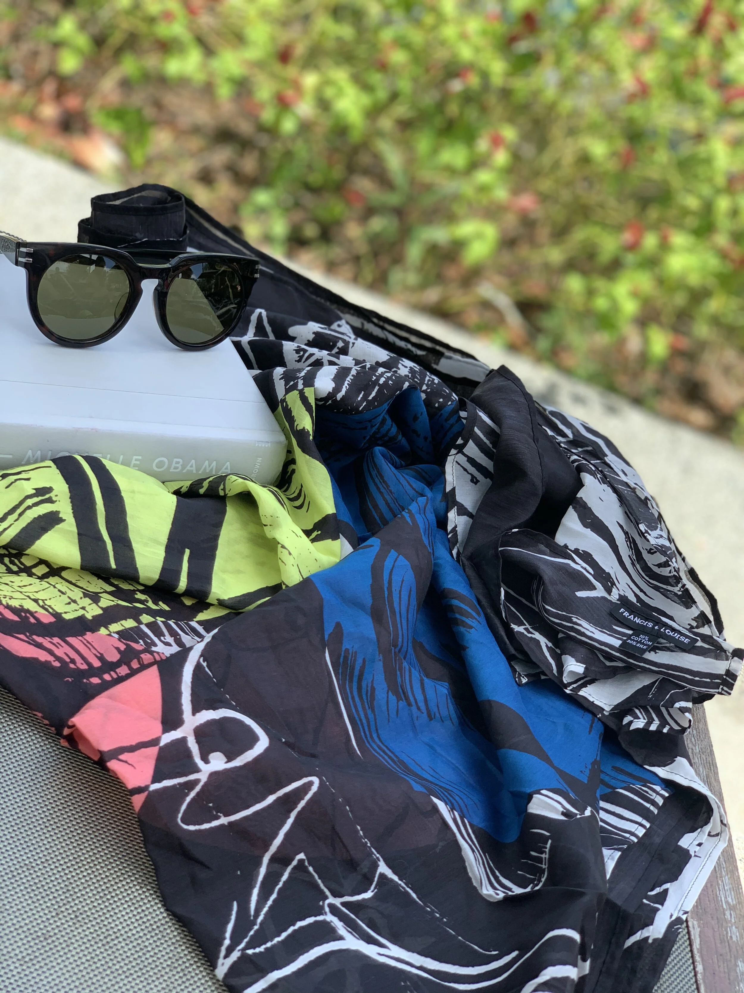 BLACK WOMEN’S COTTON SILK SCARF WITH MULTI-COLOURED ABSTRACT PATTERN OUTSIDE WITH A BOOK AND SOME SUNGLASSES.
