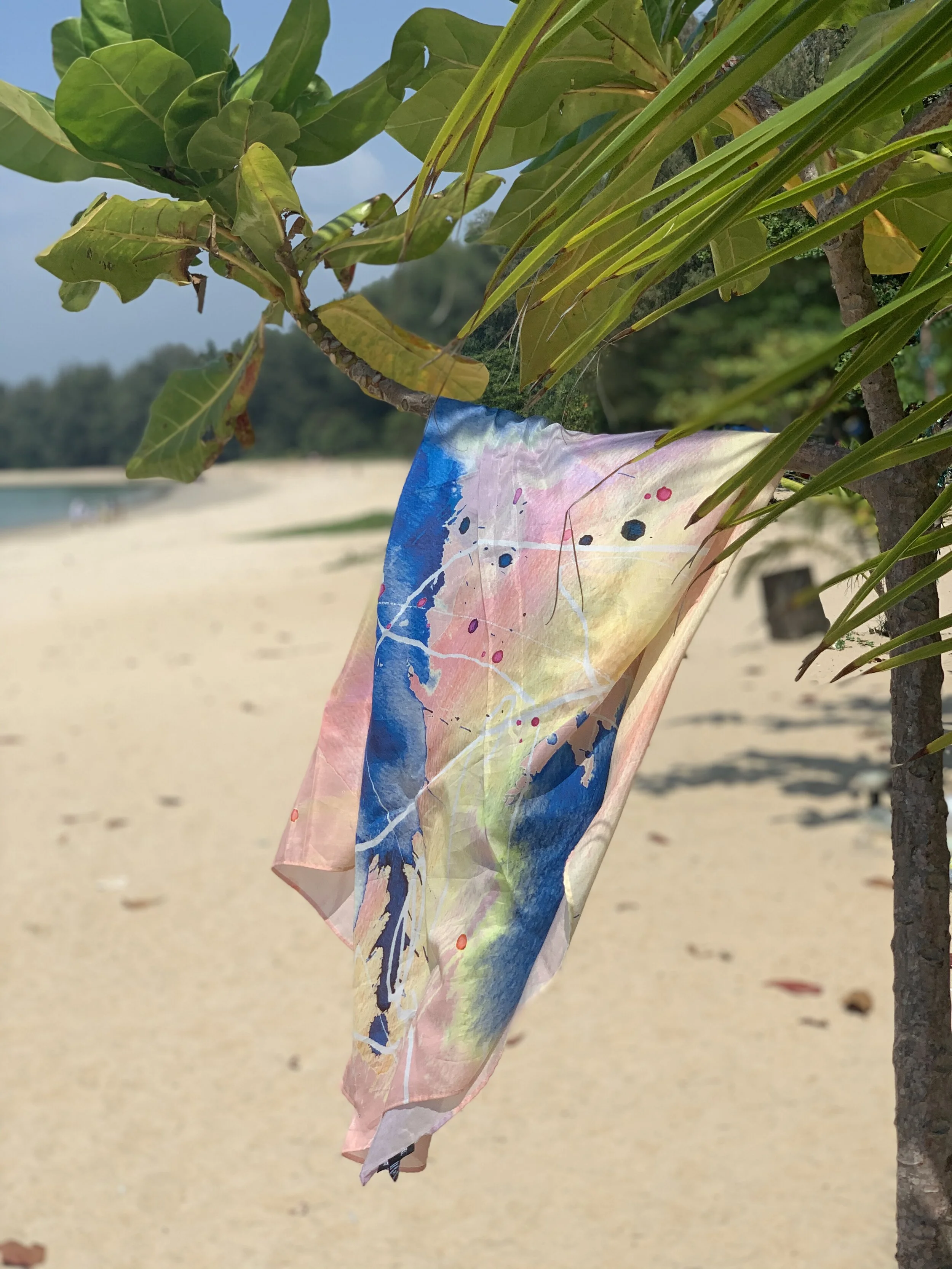 COTTON SILK WOMEN’S SQUARE SCARF WITH ABSTRACT PAINTERLY PATTERN IN PINK, BLUE AND YELLOW HANGING OVER THE BRANCH OF A TREE ON THE BEACH.. 