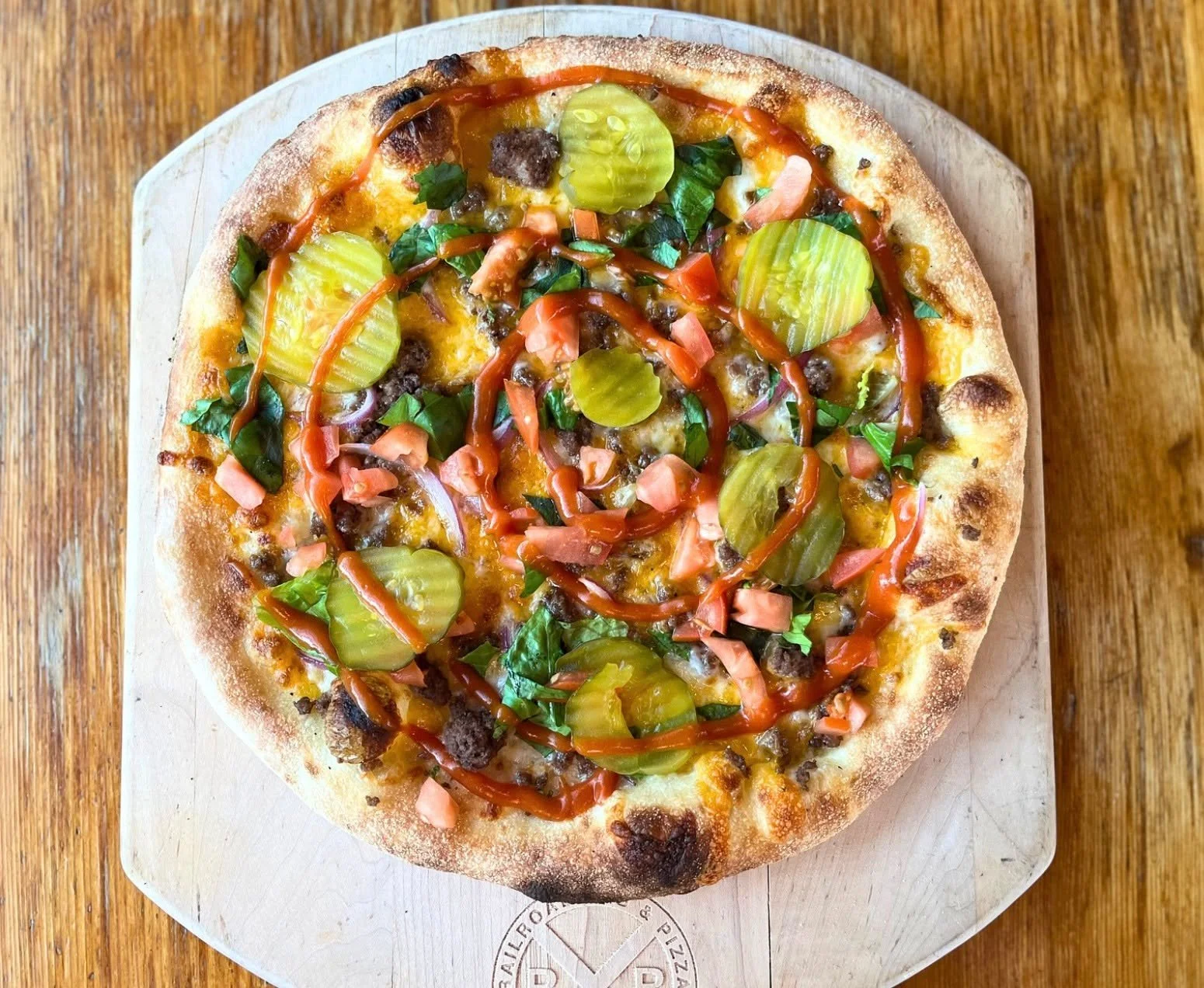 Cheeseburger Pizza is BACK! Jack Mountain Meat&rsquo;s ground beef with a blend of cheddar &amp; mozzarella cheeses, baked to perfection and topped with lettuce, diced tomatoes, red onion, and dill pickle chips. Finished with our house made burger sa
