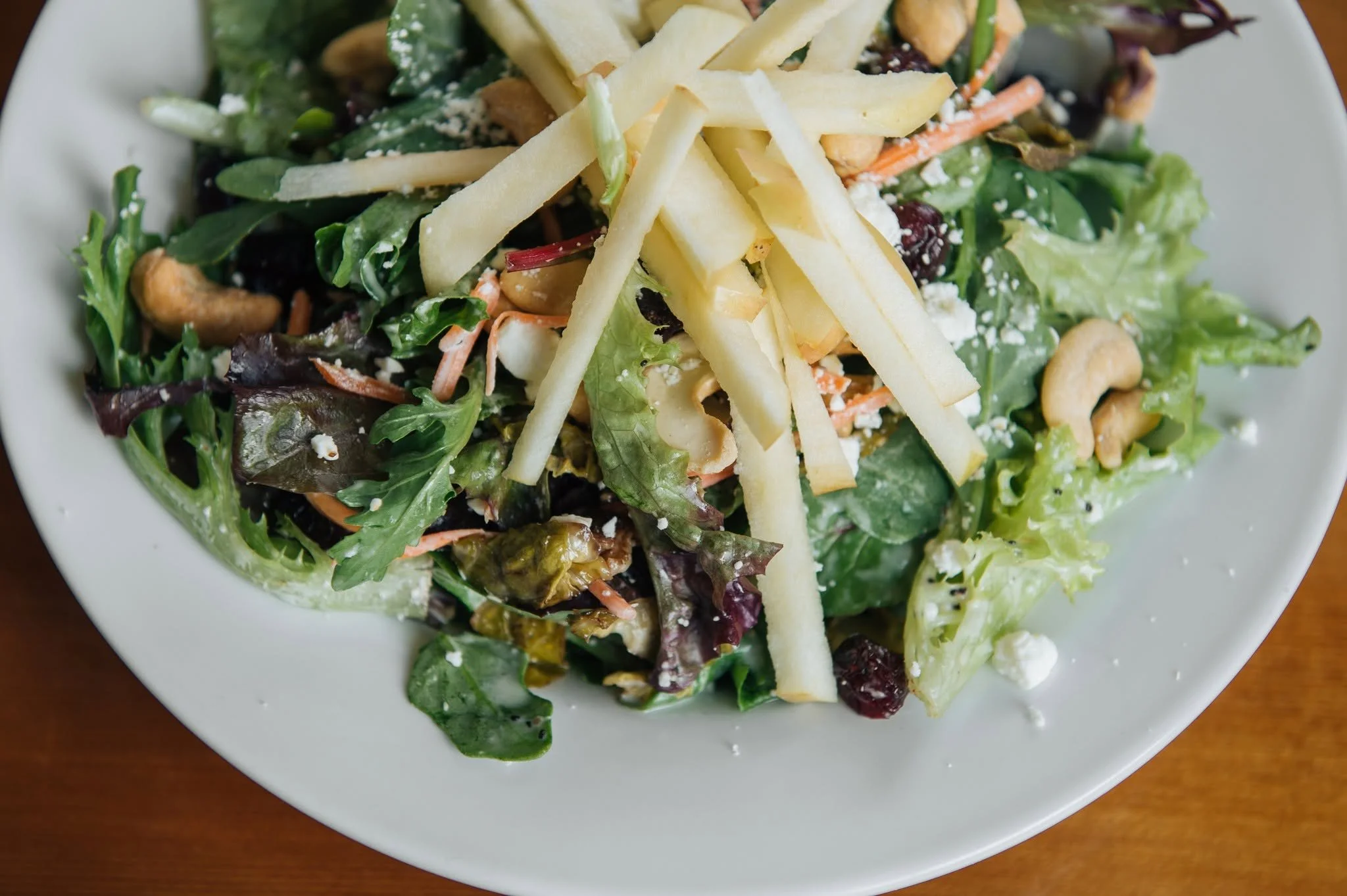 A summer favorite on the menu! Cranberry Cashew Salad is made with arugula, spring mix, brussels sprouts, carrots, basil, Craisins, and feta tossed with honey mustard dressing and topped with crisp apples and cashews.