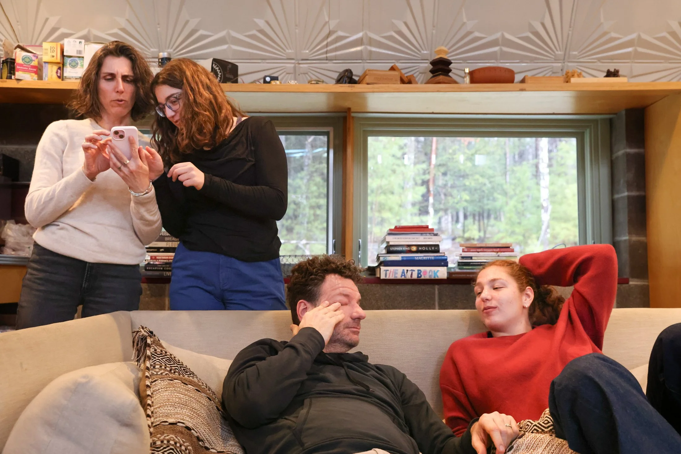 a father wipes his eye while sitting and conversing on a couch with teenage daughter; a mother and 2nd teenage daughter stand behind the couch while looking over a cellphone