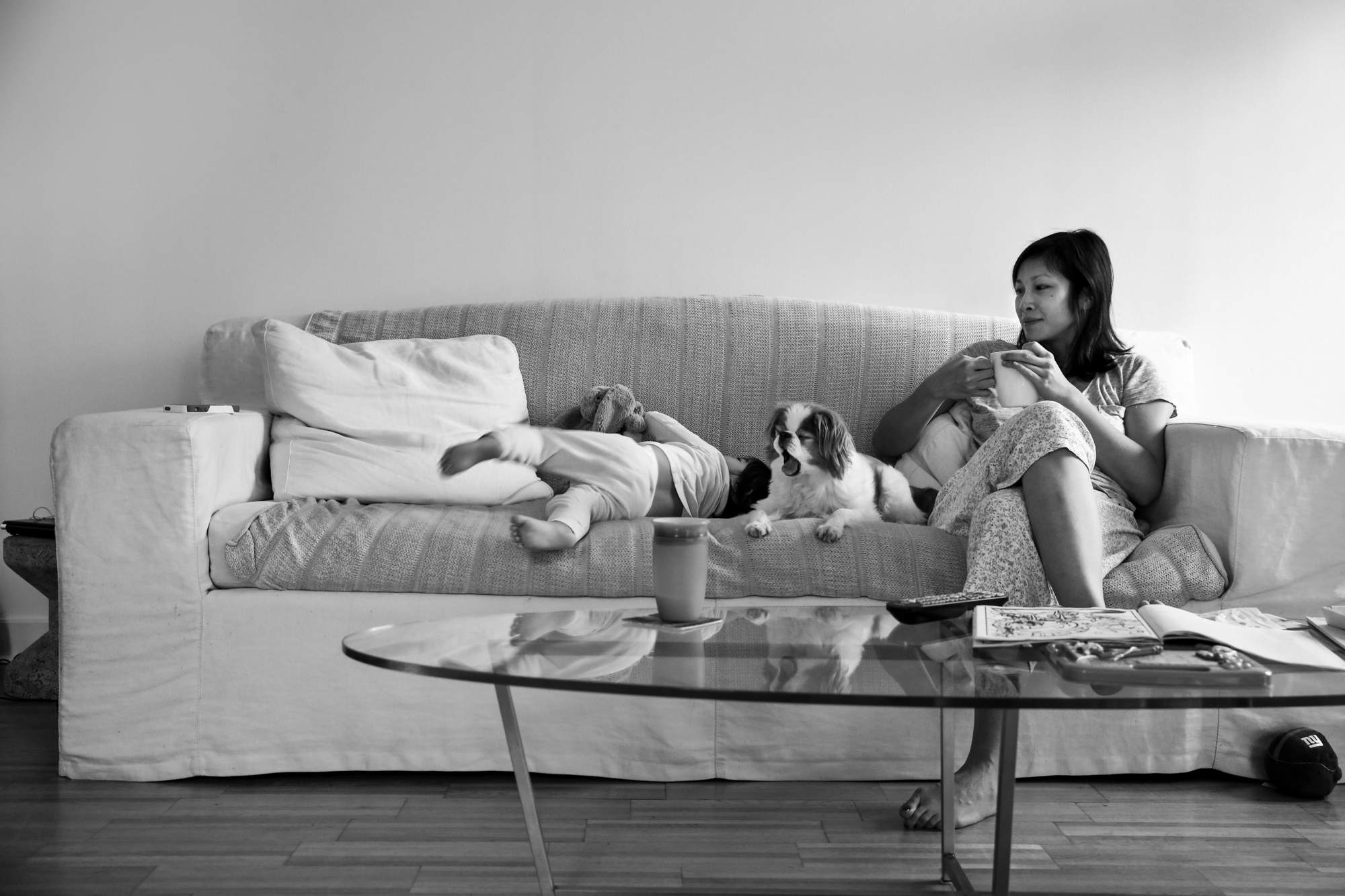 Woman with coffee sits on a couch with a girl and dog yawning