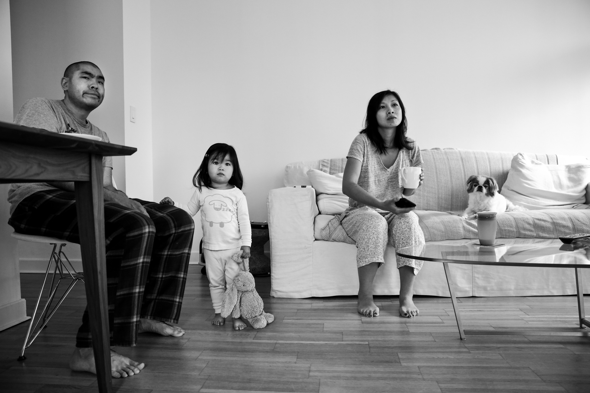 Man sits at table while child stands with stuffed bunny and woman sits on couch with dog