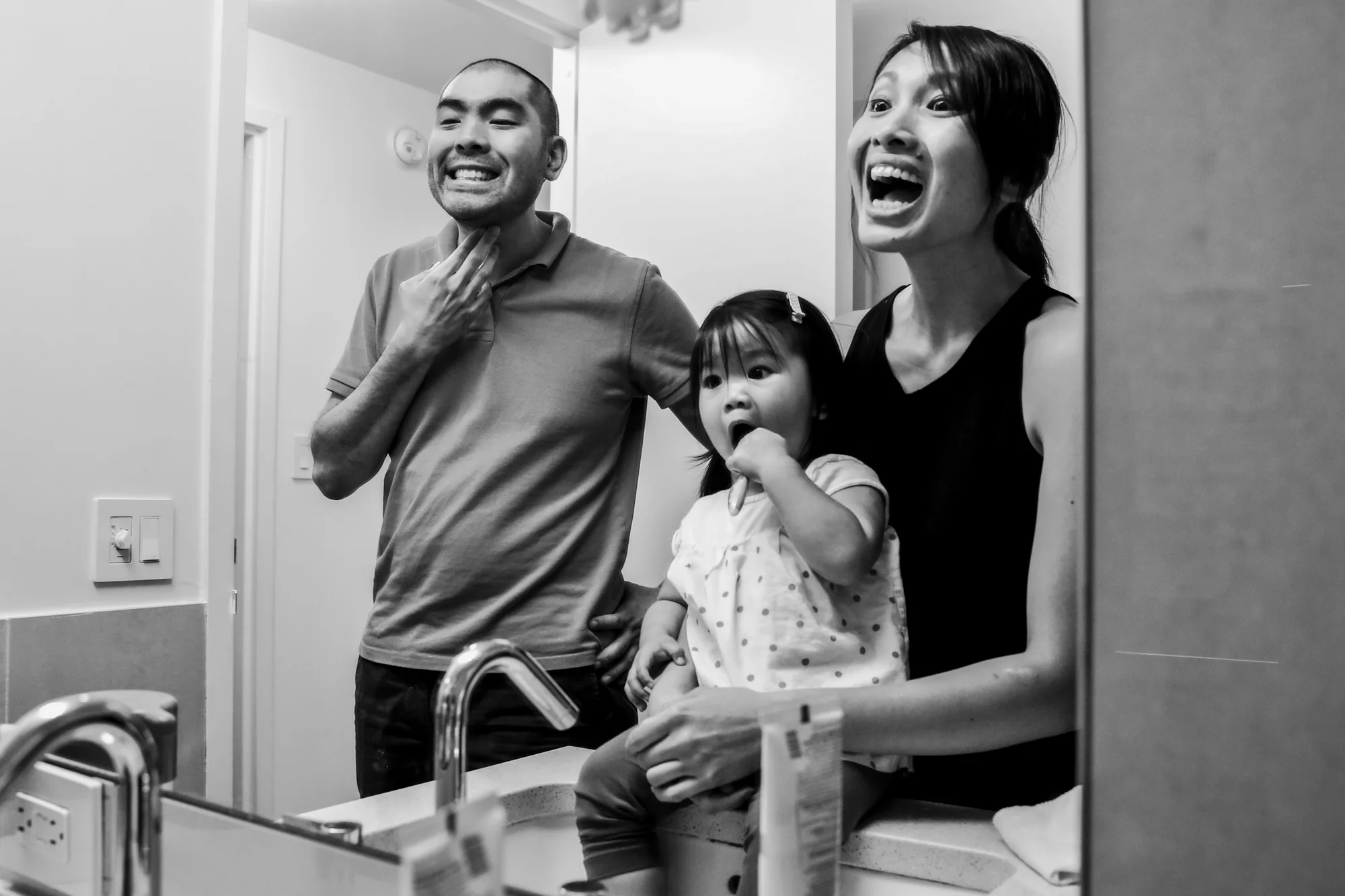 Man and woman make silly faces in the mirror while girl brushes teeth