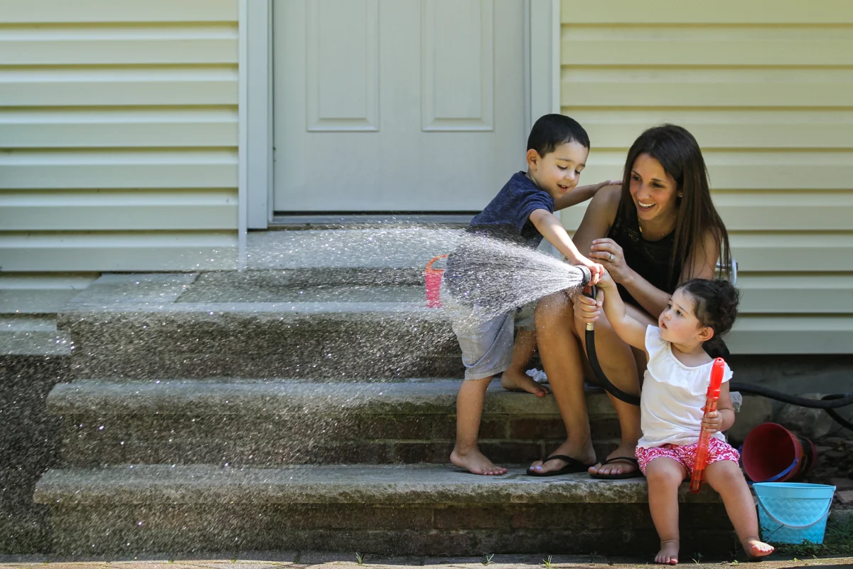 A boy and girl help their mom spray a garden hose while sitting on steps