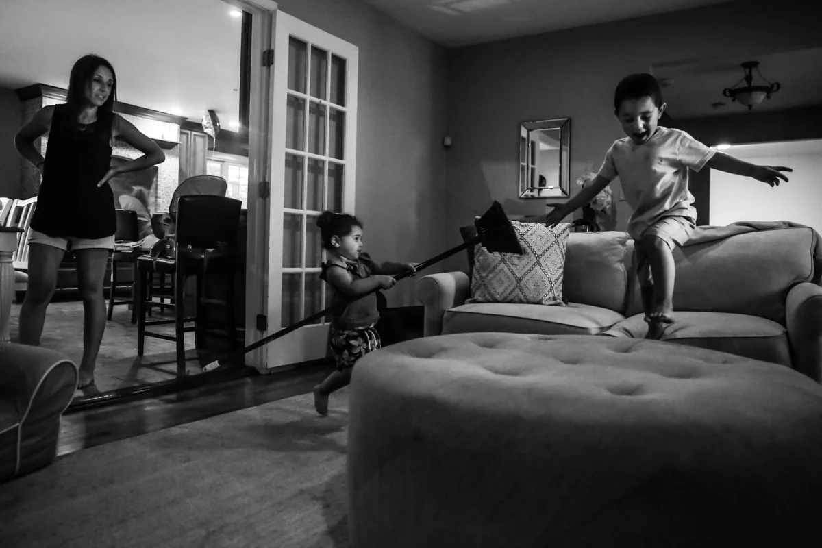 Boy jumps from couch to ottoman while girl chases him with a broom and woman watches from the side