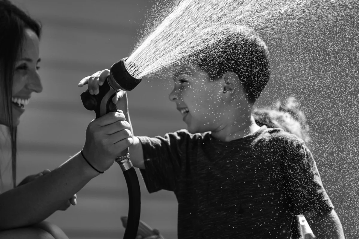 Mother and son smile at each other while spraying a garden hose