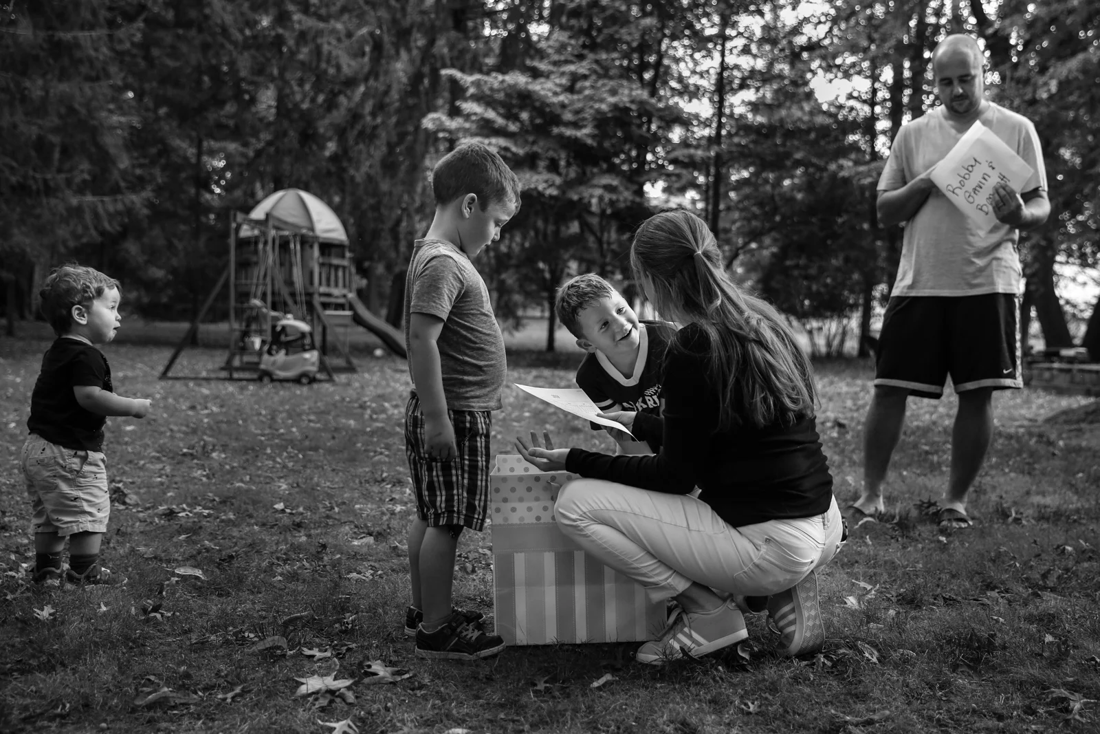 Woman squats and holds paper in hand while boy looks amusingly at her, Another boy looks down at paper. Another boy stands looking surprised at scene. Man looks through contents of envelope