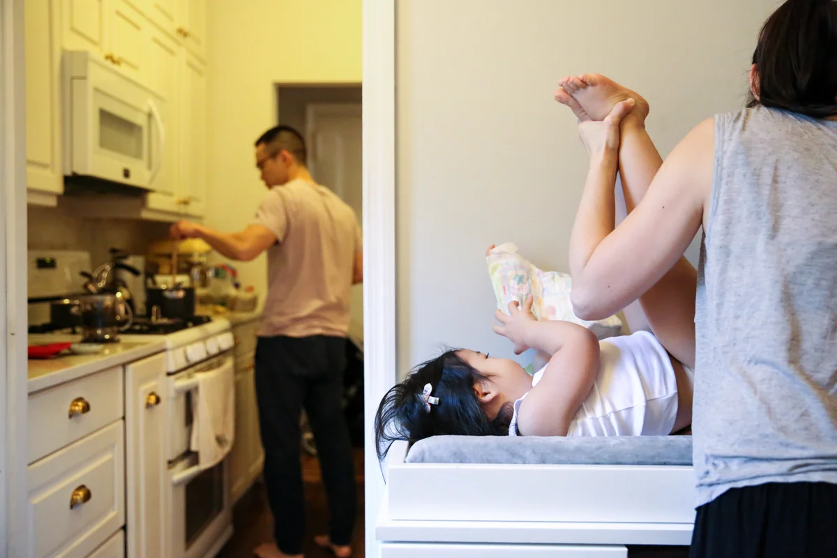 Woman changes diaper of girl while man cooks breakfast