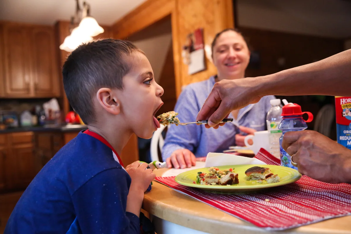 Boy opens wide for a forkful of eggs while woman smiles in background