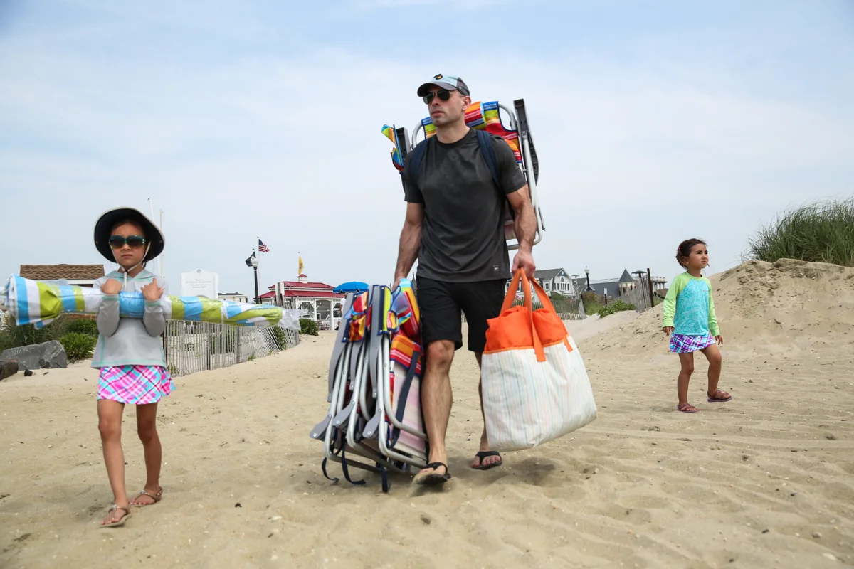Man walks with 2 children while carrying beach chairs and beach bag onto the sandy beach. Girl with hat and sunglasses carries beach umbrella
