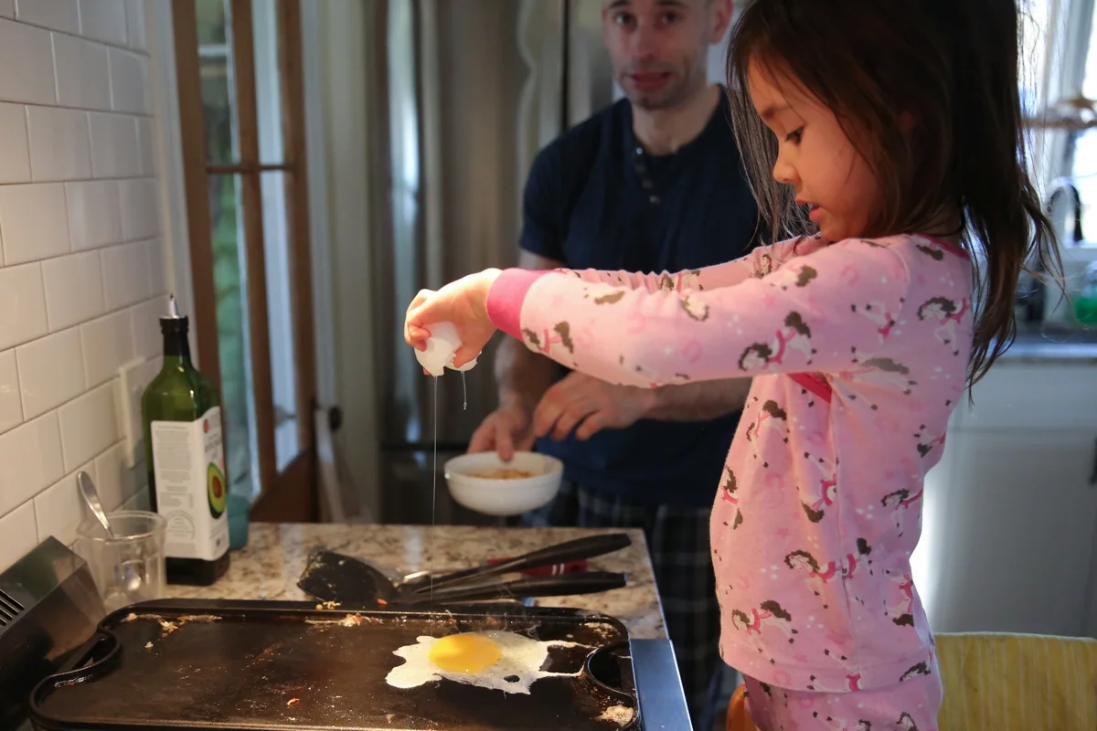 Girl cracks egg on a stovetop griddle while man looks apprehensively