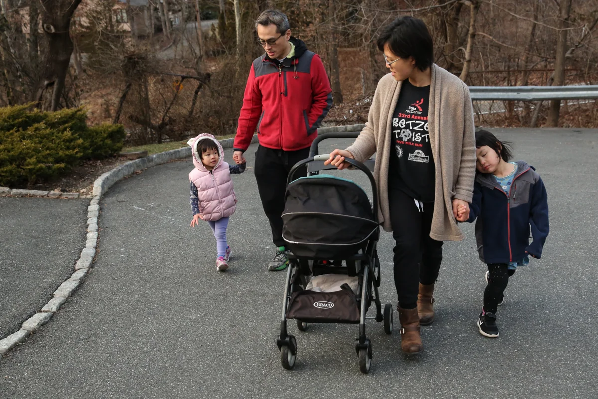 Man, woman and two children walk on street while woman pushes snap n go stroller. Girl holds woman’s hand while eyes closed