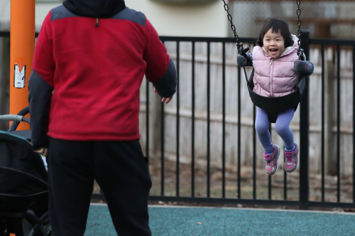 Man in red sweatshirt pushes girl wearing pink vest on swing