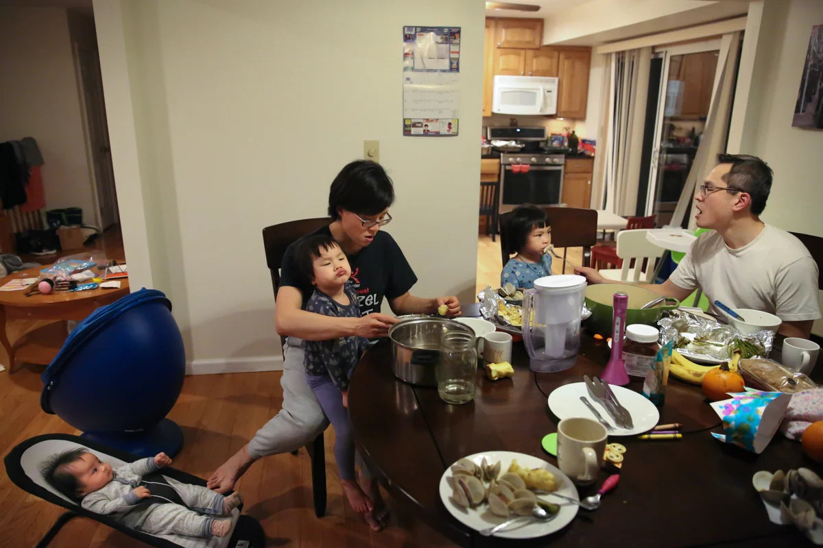 Family sits at dinner table—parents try to feed two girls while baby sits in bouncer on floor with mother pushing with her foot